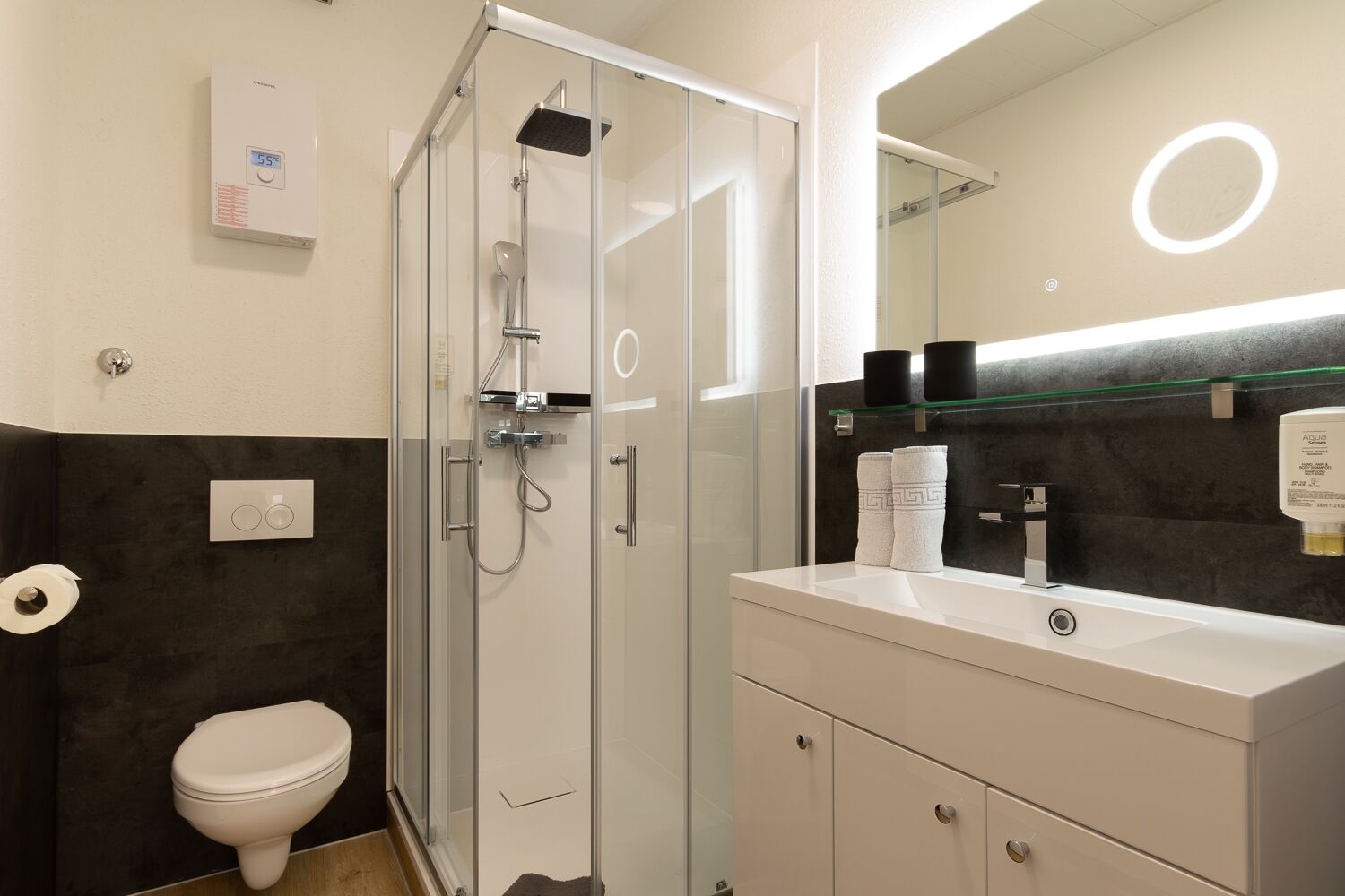 Une salle de bain moderne avec une douche à parois de verre, des toilettes blanches, un meuble-lavabo avec un miroir et un distributeur de serviettes.