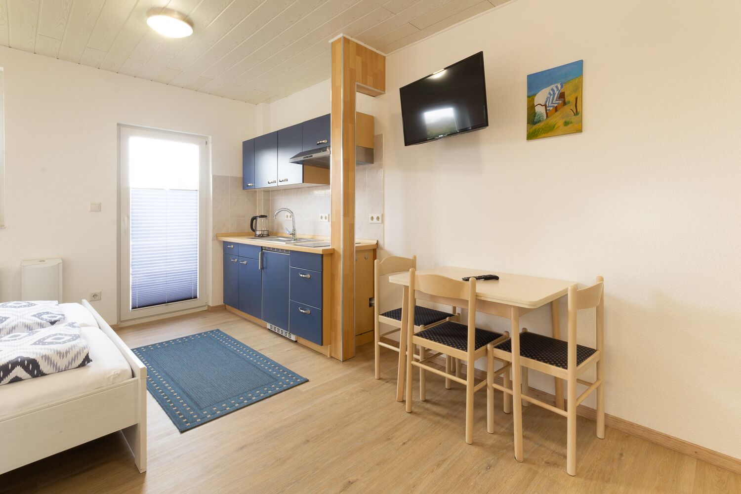 A compact room with a bed, a small kitchenette, a dining table with chairs, a TV mounted on the wall, and a window with blinds.