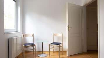 Een kamer met twee houten stoelen en een klein glazen tafeltje, een witte radiator, en een deur naar een andere kamer.