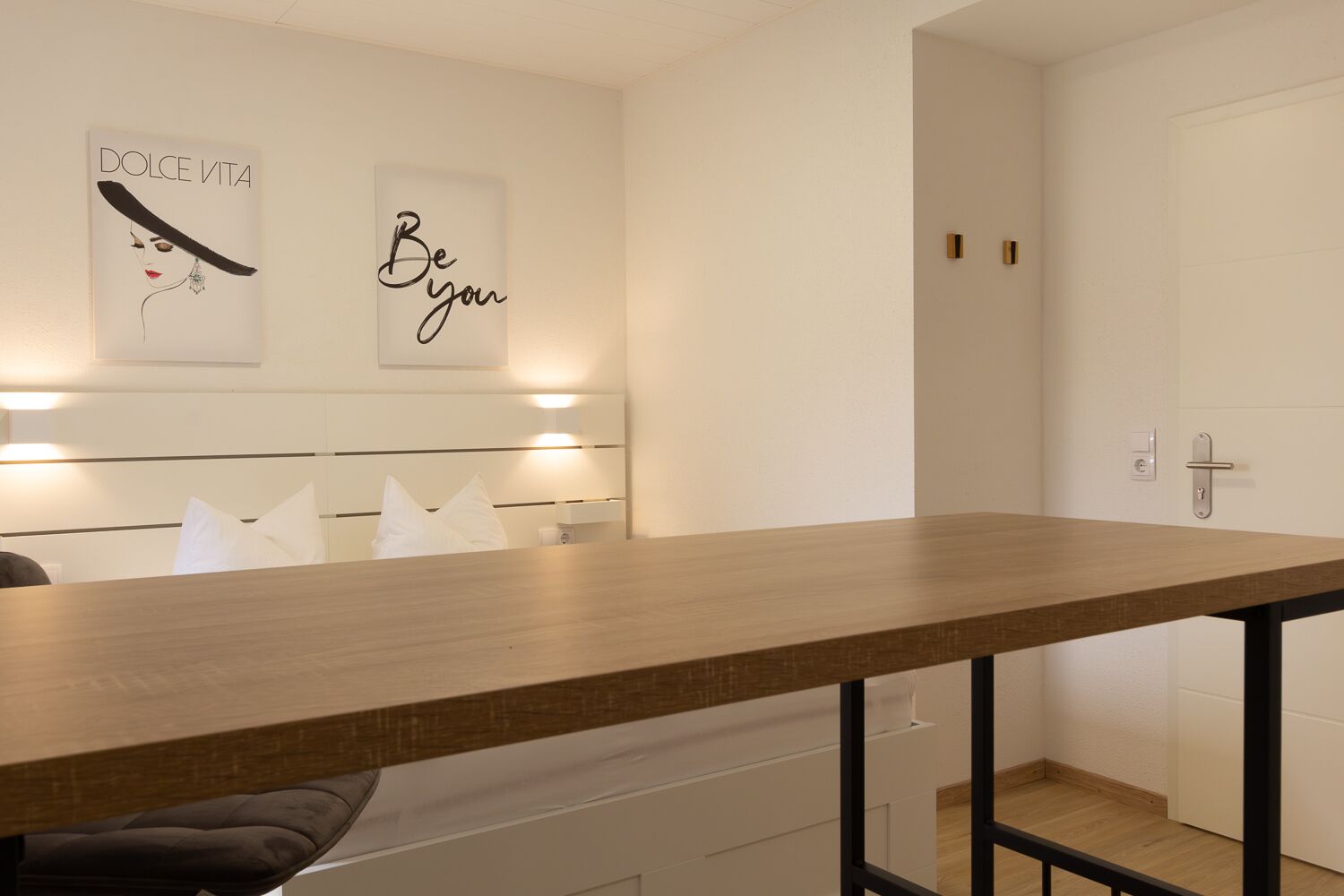 A modern hotel room with a wooden dining table, white headboard, and two wall-mounted light fixtures.