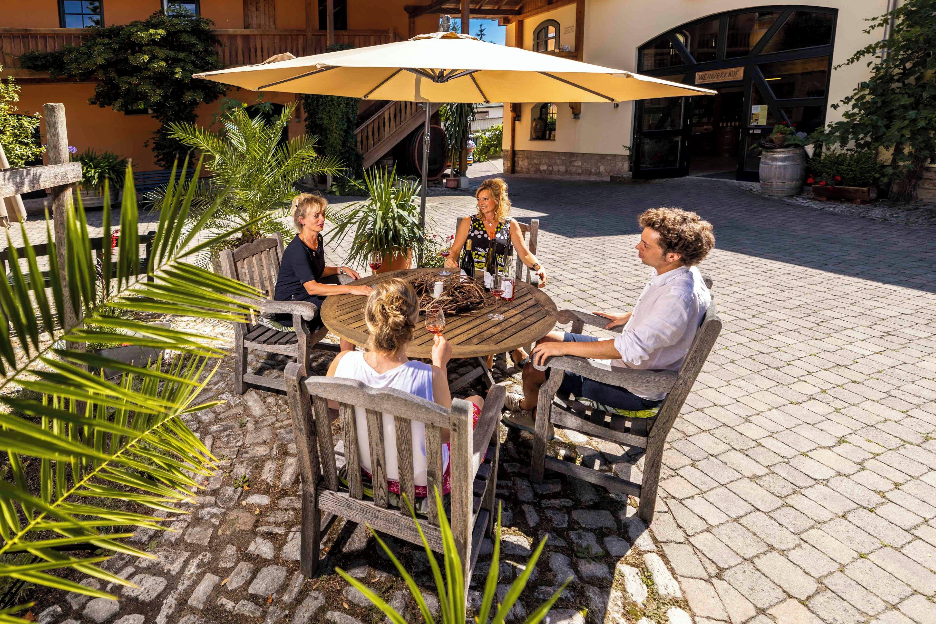 Um grupo de pessoas sentadas em uma mesa ao ar livre, sob um guarda-sol grande, cercado por plantas em vasos e um pátio de pedras irregulares.