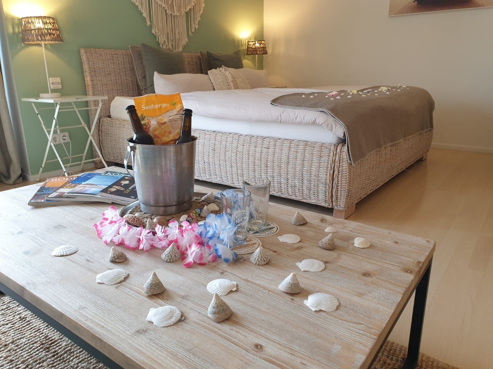 Una habitación ordenada con una cama, una mesita de café con conchas marinas y una botella de Seeburger, y una revista.