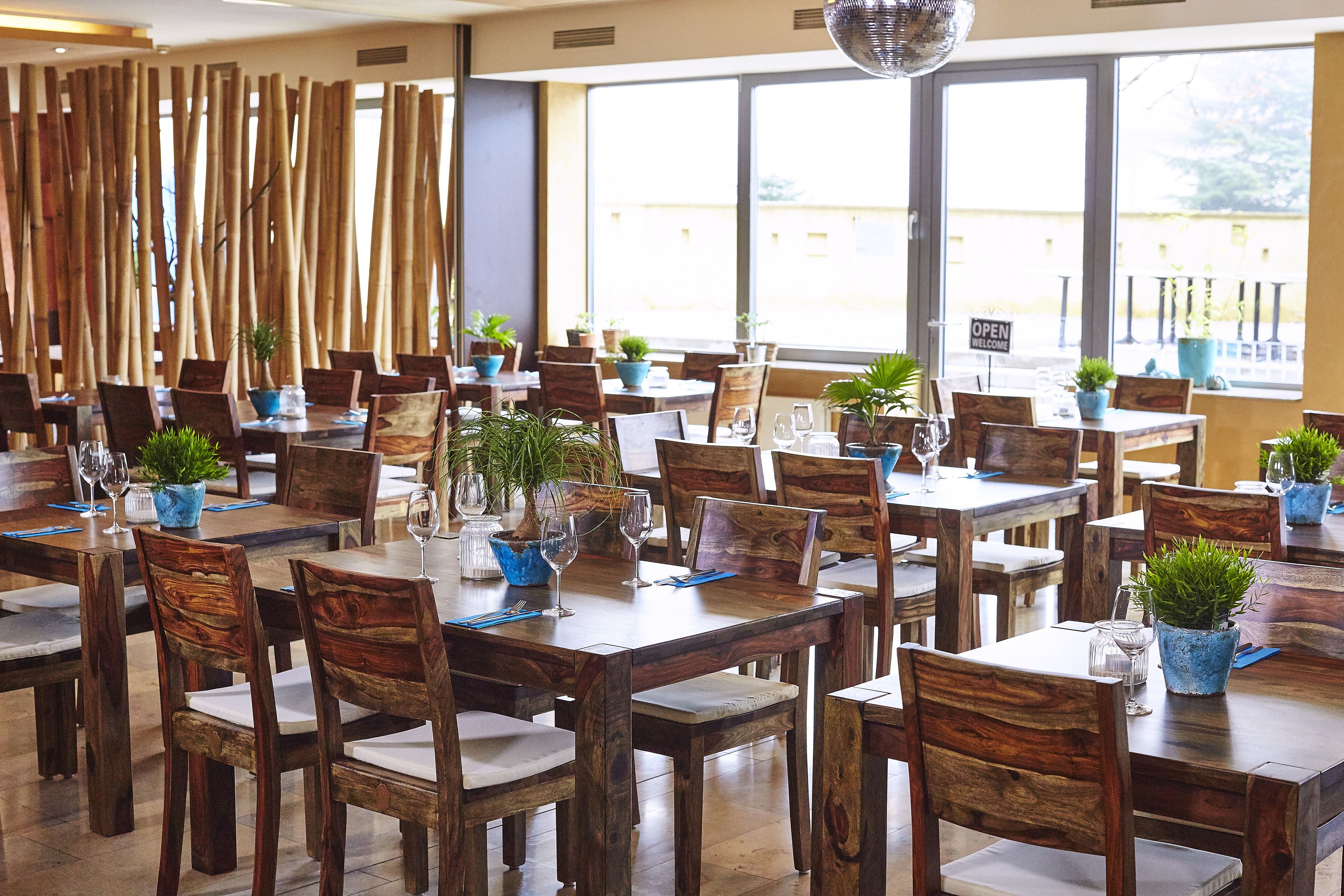 Un restaurante con mesas y sillas de madera, plantas en macetas y vistas amplias por los grandes ventanales.