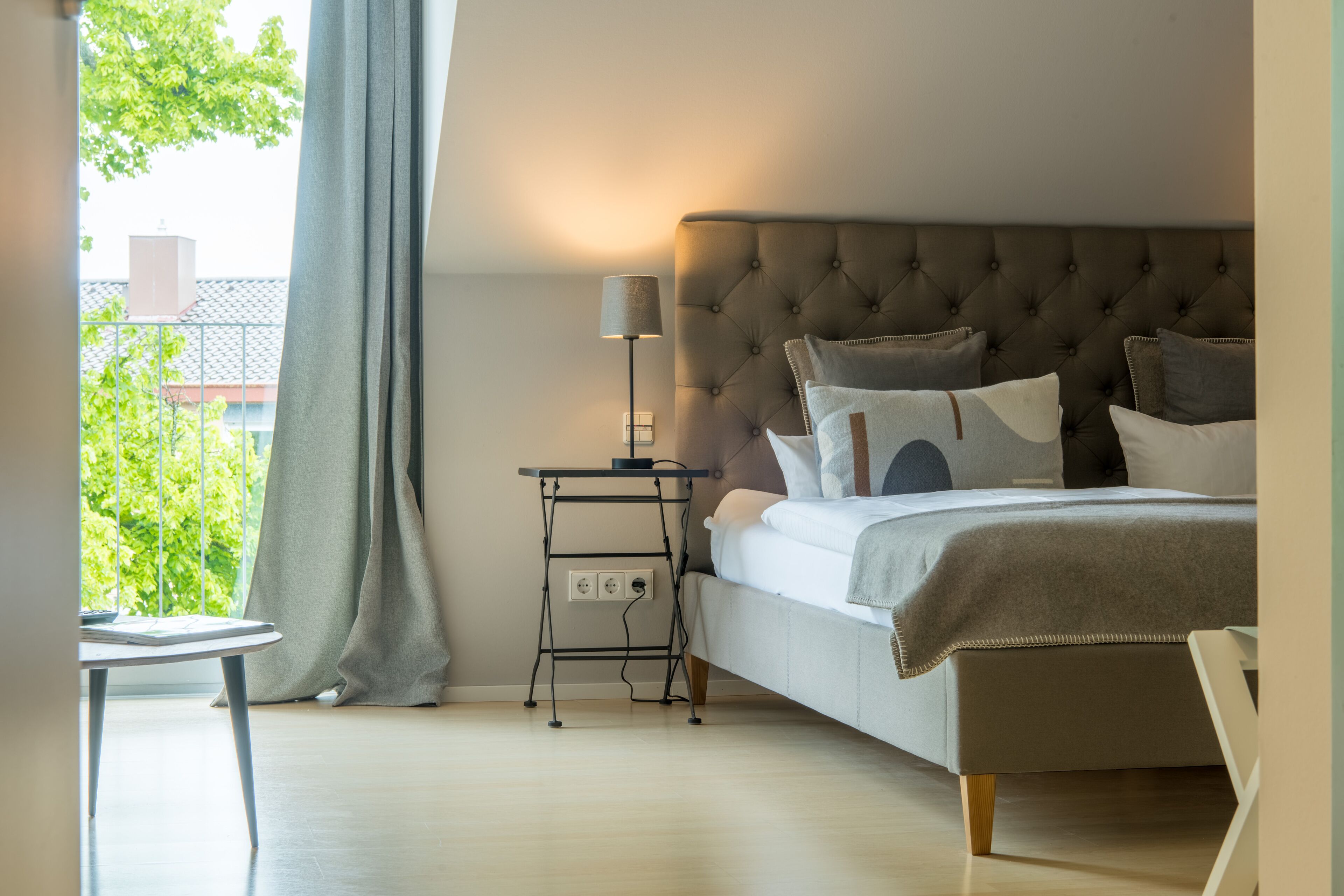 A bedroom with a bed, bedside table, lamp, and a view of greenery outside.
