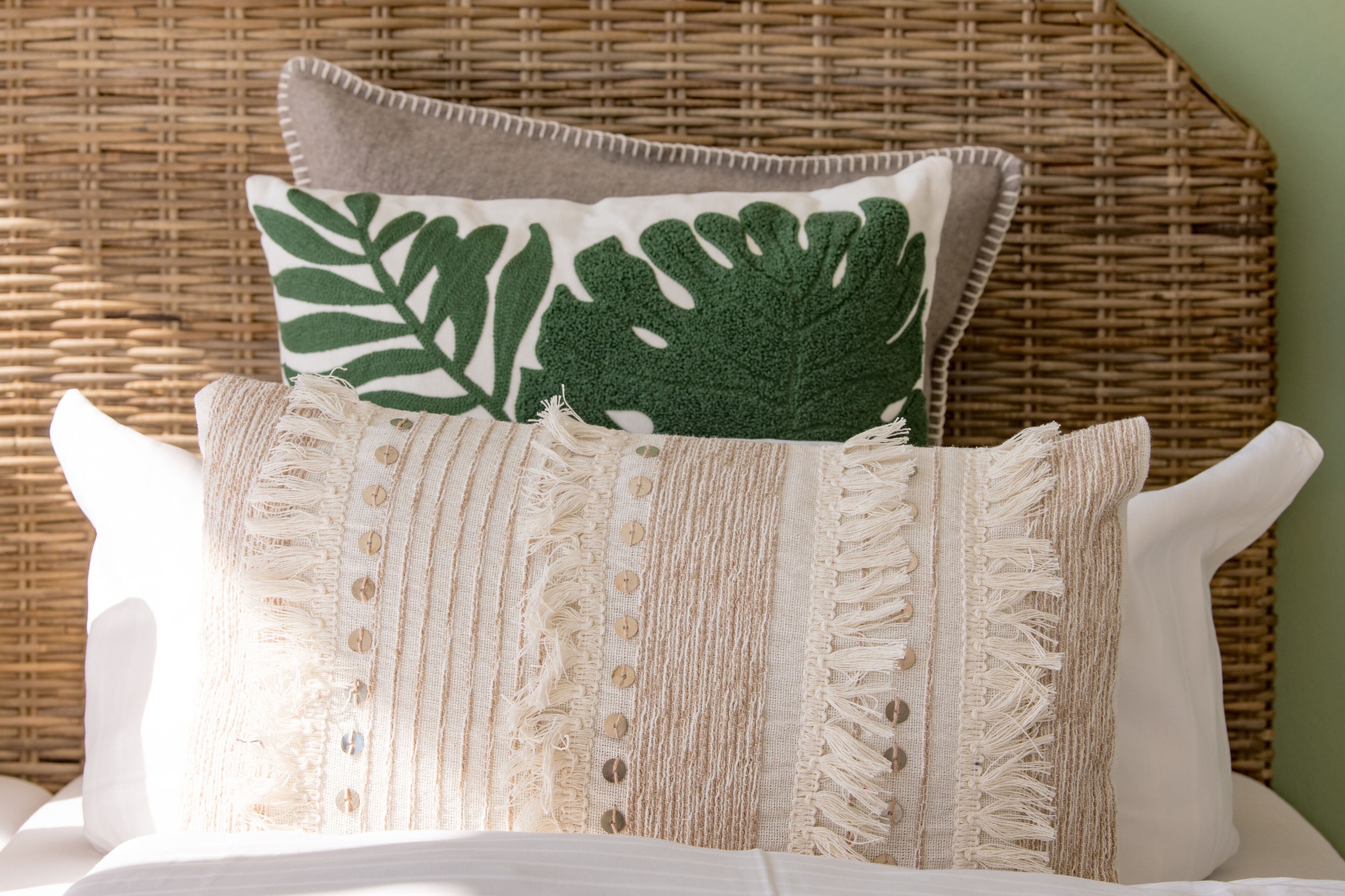 Una cama con un almohadón blanco y beige a rayas, un cojín con estampado de hojas verdes y un almohadón blanco.