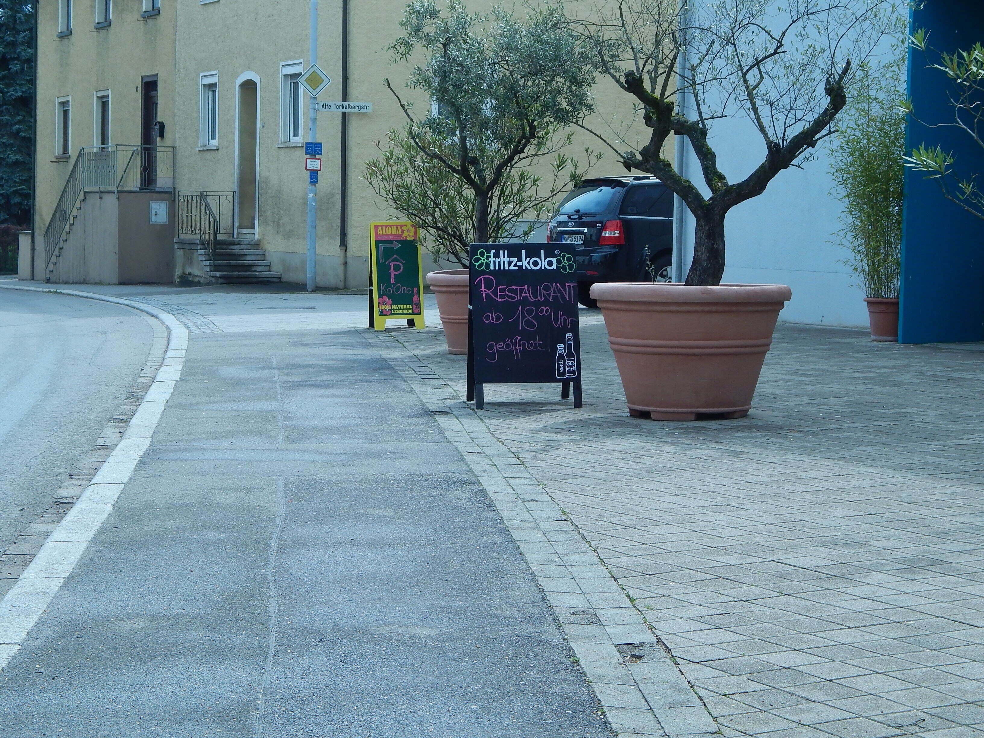 A street with a parked car, a signboard for Fritz-Kola Restaurant, and large potted plants.