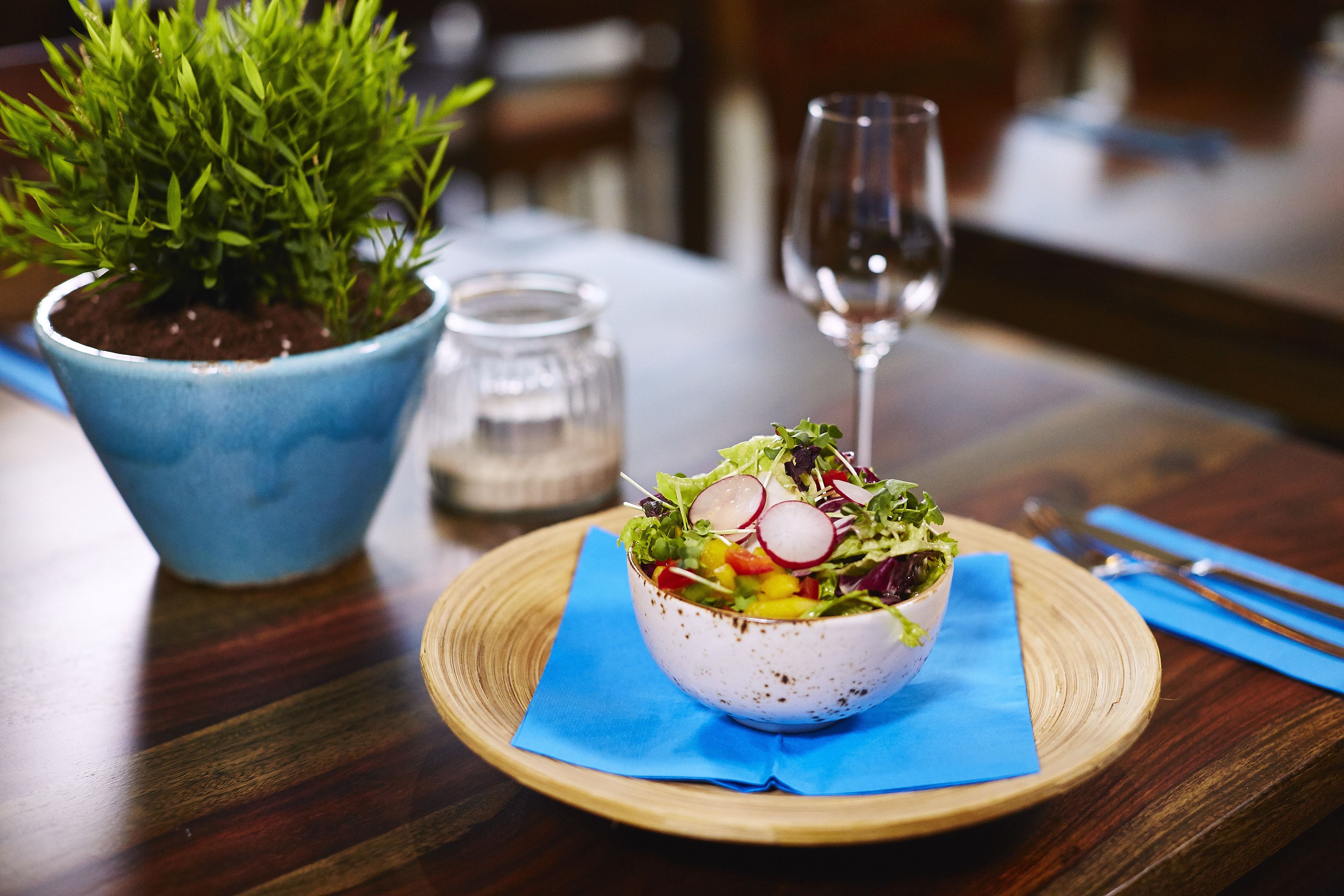 Uma salada numa tigela sobre uma mesa de madeira, com um guardanapo azul, um copo de vinho e um vaso de planta ao fundo.
