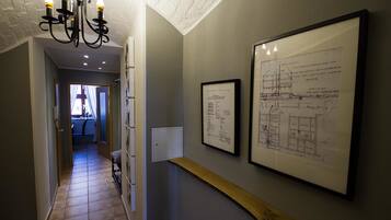 A hallway with a chandelier, framed architectural drawings on the wall, and a wooden shelf.