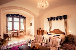 Une chambre d’hôtel avec un grand lit, deux fauteuils, un bureau, un lustre et une table de chevet.