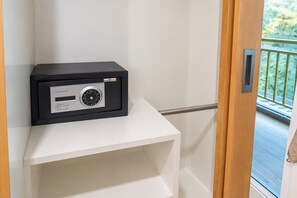 A black safe with operating instructions on a white shelf in a room with a wooden door and a balcony view. - T Sleep Place (Pattaya)