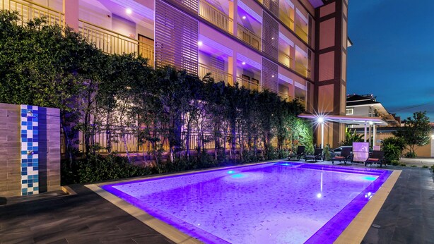 Uma piscina bem iluminada com um deck de azulejos, cercada por vegetação exuberante e um edifício com várias andares e varandas.