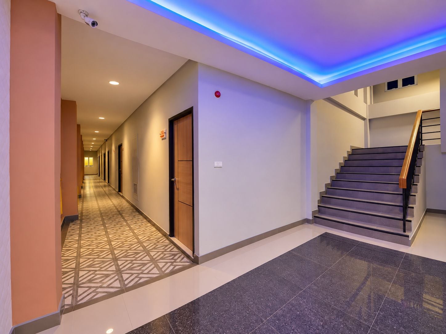 A hotel corridor with a patterned floor, a staircase, and a security camera.