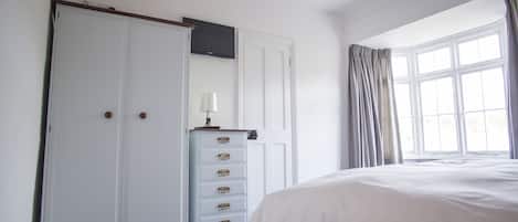 Ein Schlafzimmer mit einem großen Bett, einem Kleiderschrank, einem Schminktisch und einem Fernseher, sowie einem Fenster mit Vorhängen.
