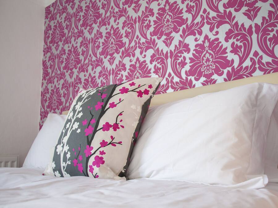 Cama con ropa blanca y cabecera frente a un papel tapiz floral rosa.