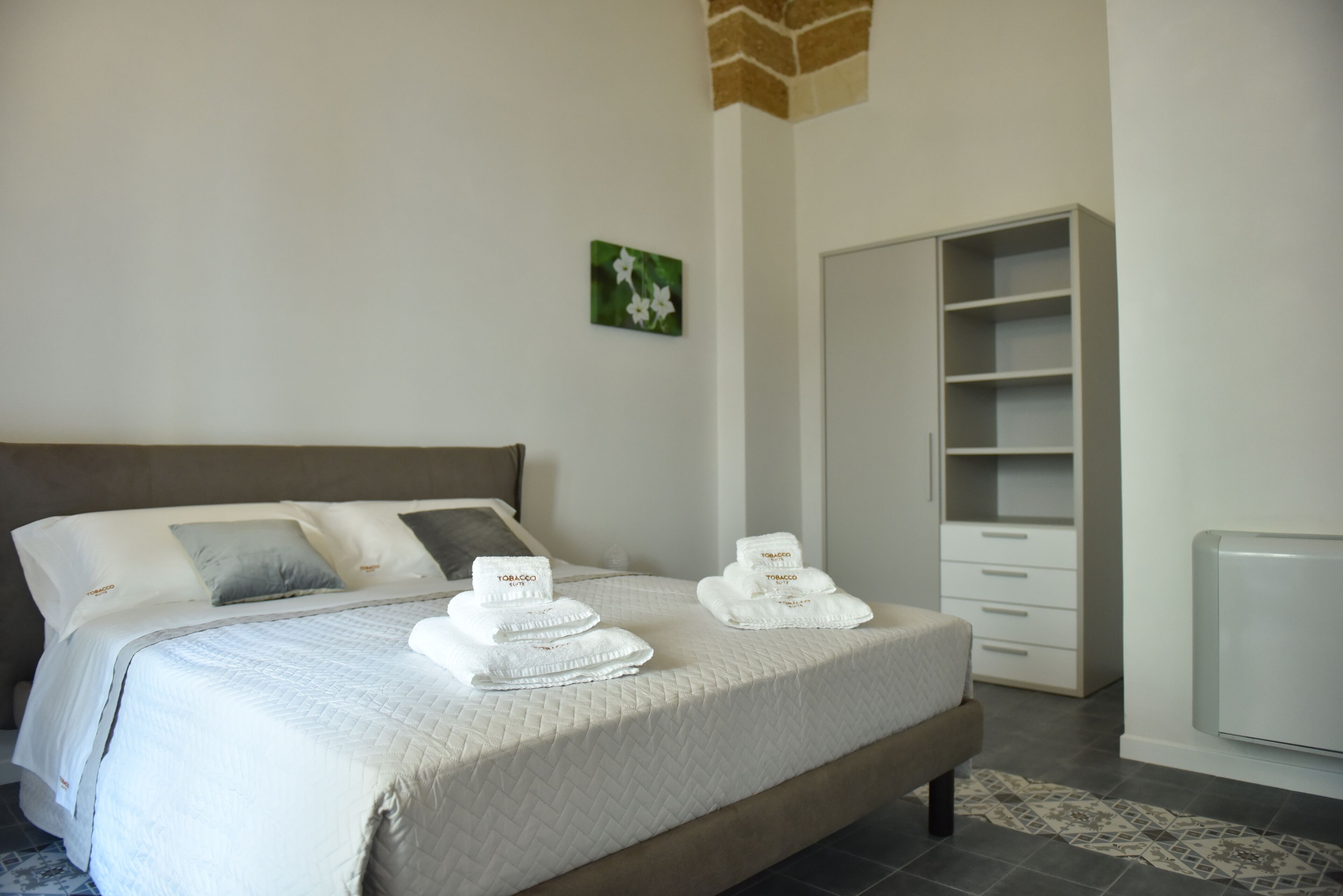 A neatly made bed with white linens and towels, a grey headboard, a small green floral artwork on the wall, and a white wardrobe with drawers.