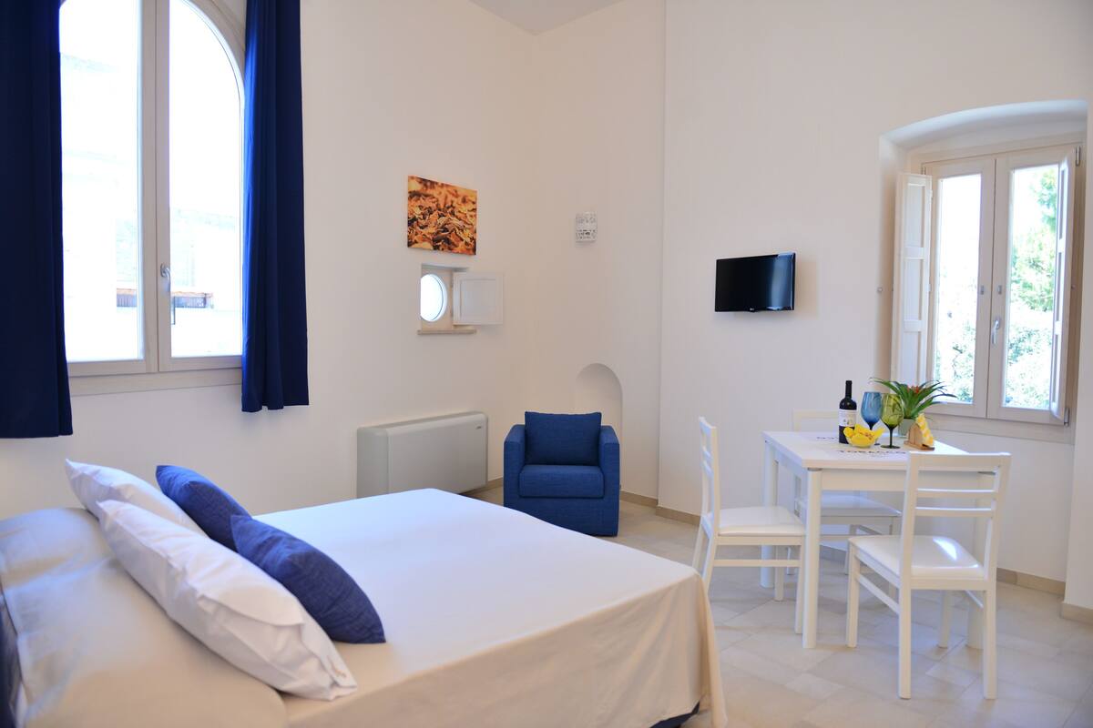 Quarto de hotel com cama, escrivaninha com cadeira, televisão e janela com cortinas.