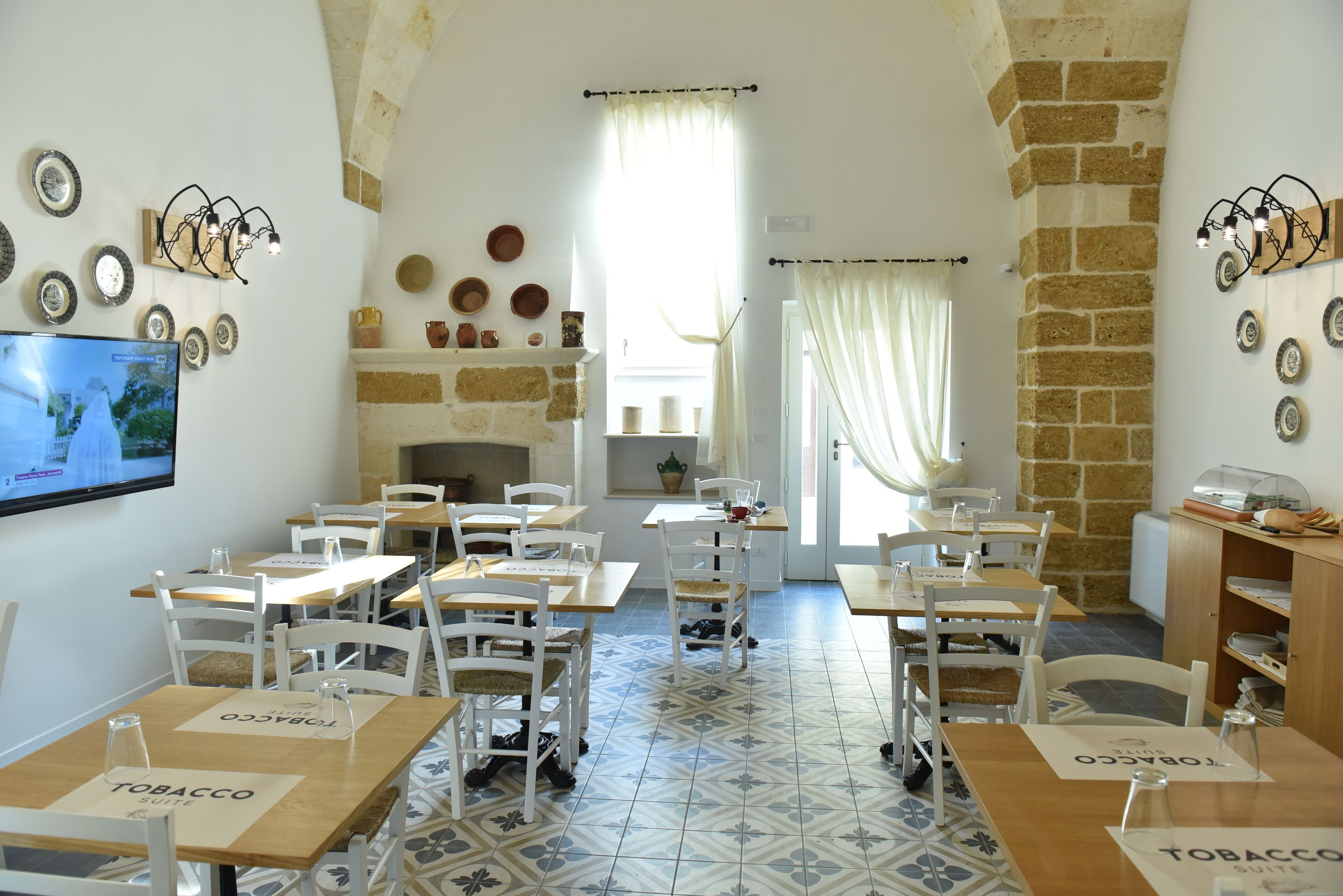 A dining area with tables and chairs, a television on the wall, and a stone wall with decorative plates.