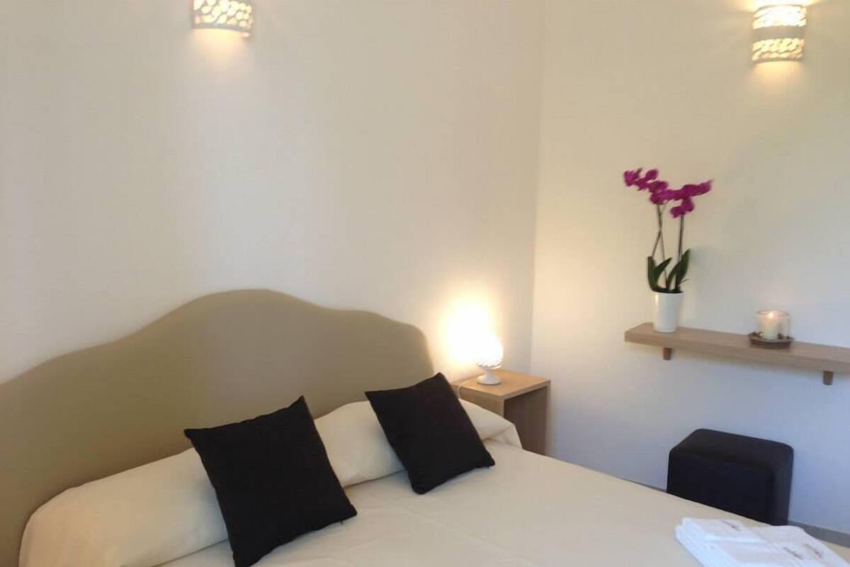 Cama bem arrumada com cabeceira, dois travesseiros pretos, abajur, pequena estante com um vaso de flores e toalha dobrada na mesinha de cabeceira.
