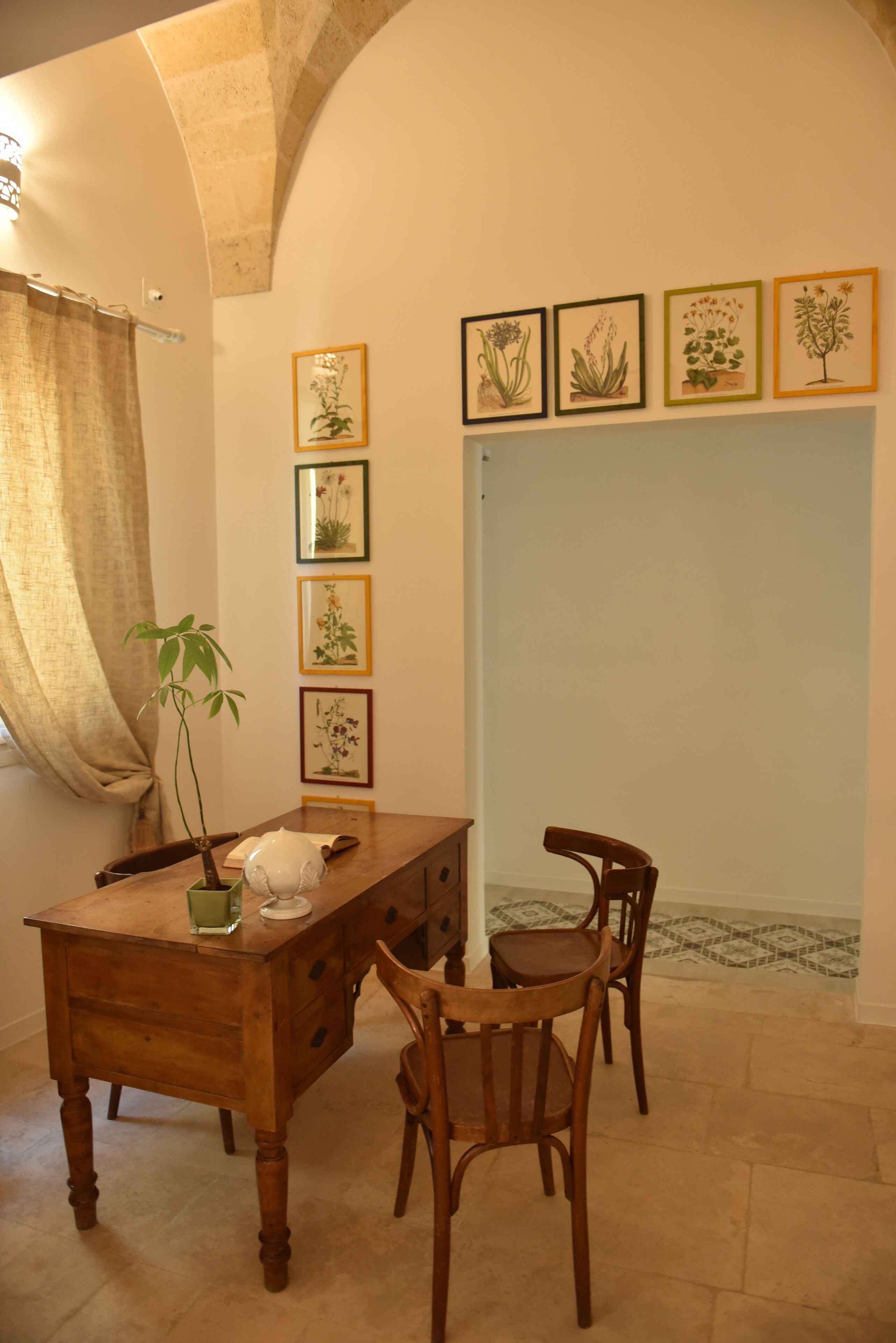 A dining area with a wooden table, chairs, a potted plant, and framed botanical illustrations on the wall.