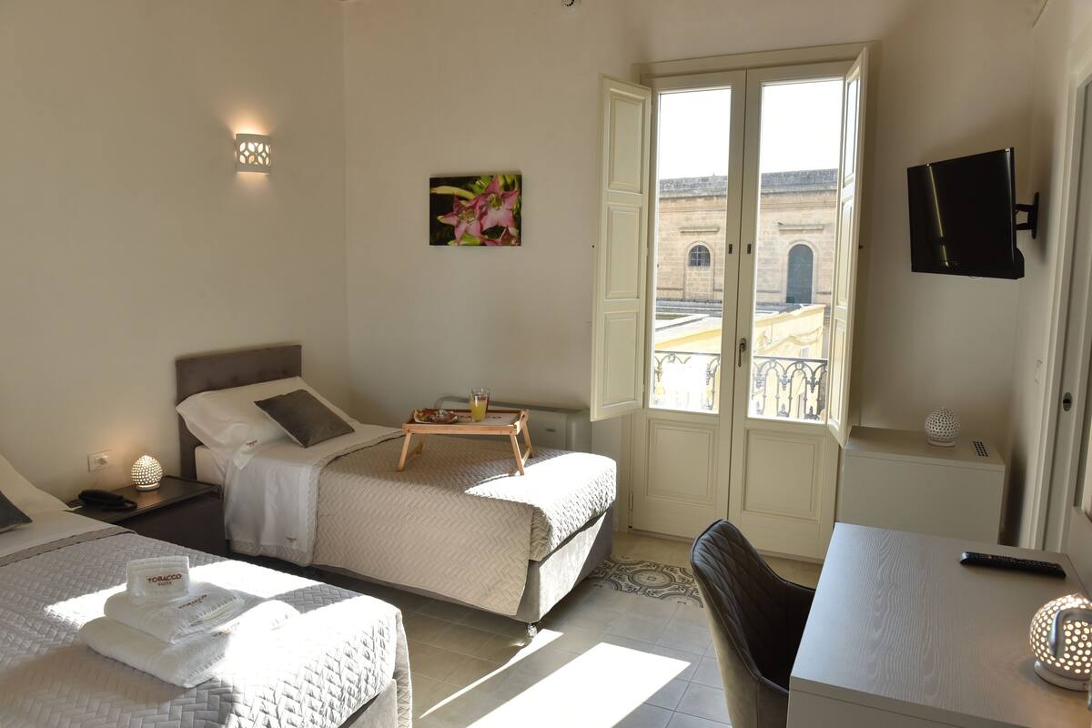 Quarto de hotel com duas camas, uma escrivaninha, uma cadeira, televisão e varanda com vista para a cidade.