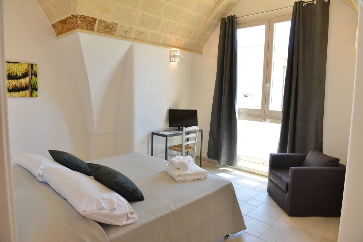 Quarto com cama, poltrona, televisão e janela com cortinas.