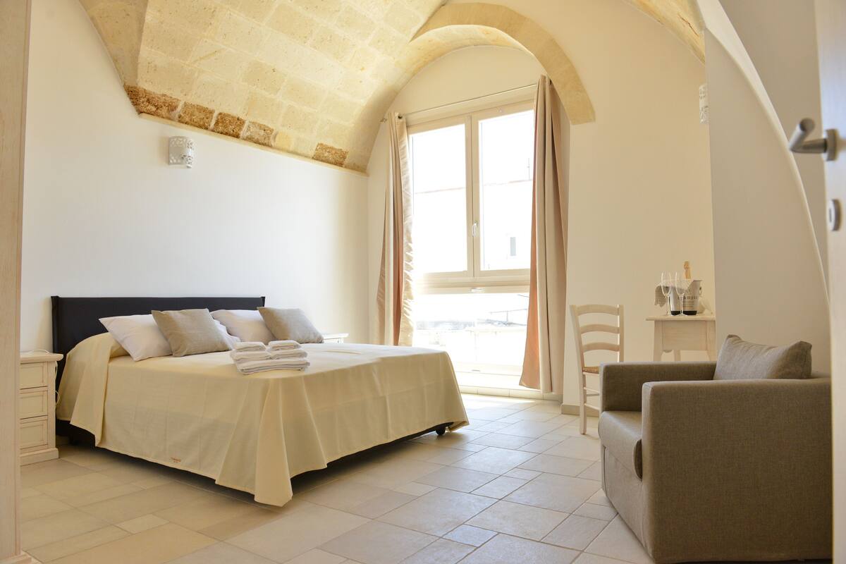 Quarto com cama, cadeira, mesa de cabeceira e janela com cortinas.