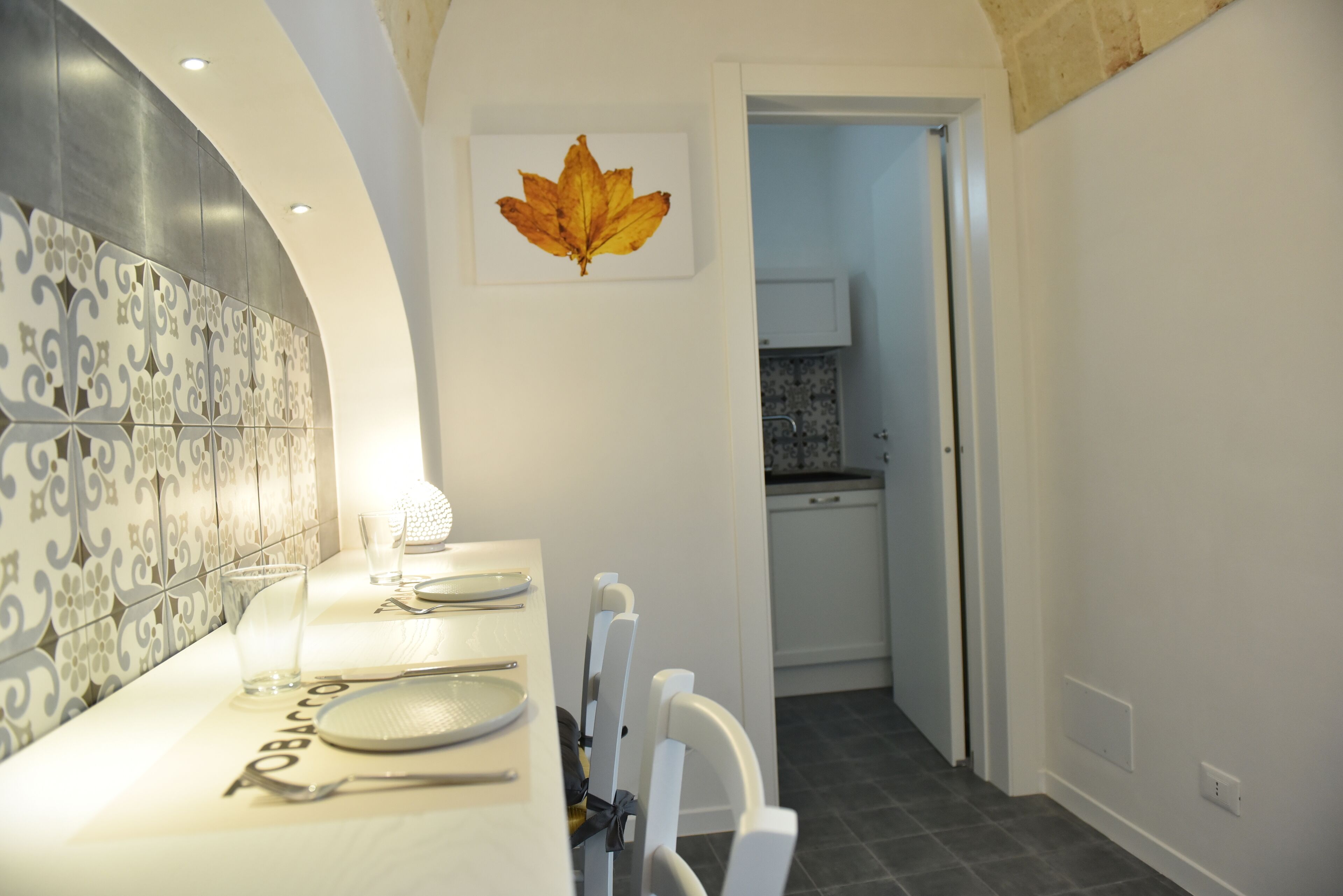 Una cocina moderna con armarios blancos, un comedor con una mesa blanca dispuesta para cuatro personas y una obra de arte decorativa con hojas en la pared.