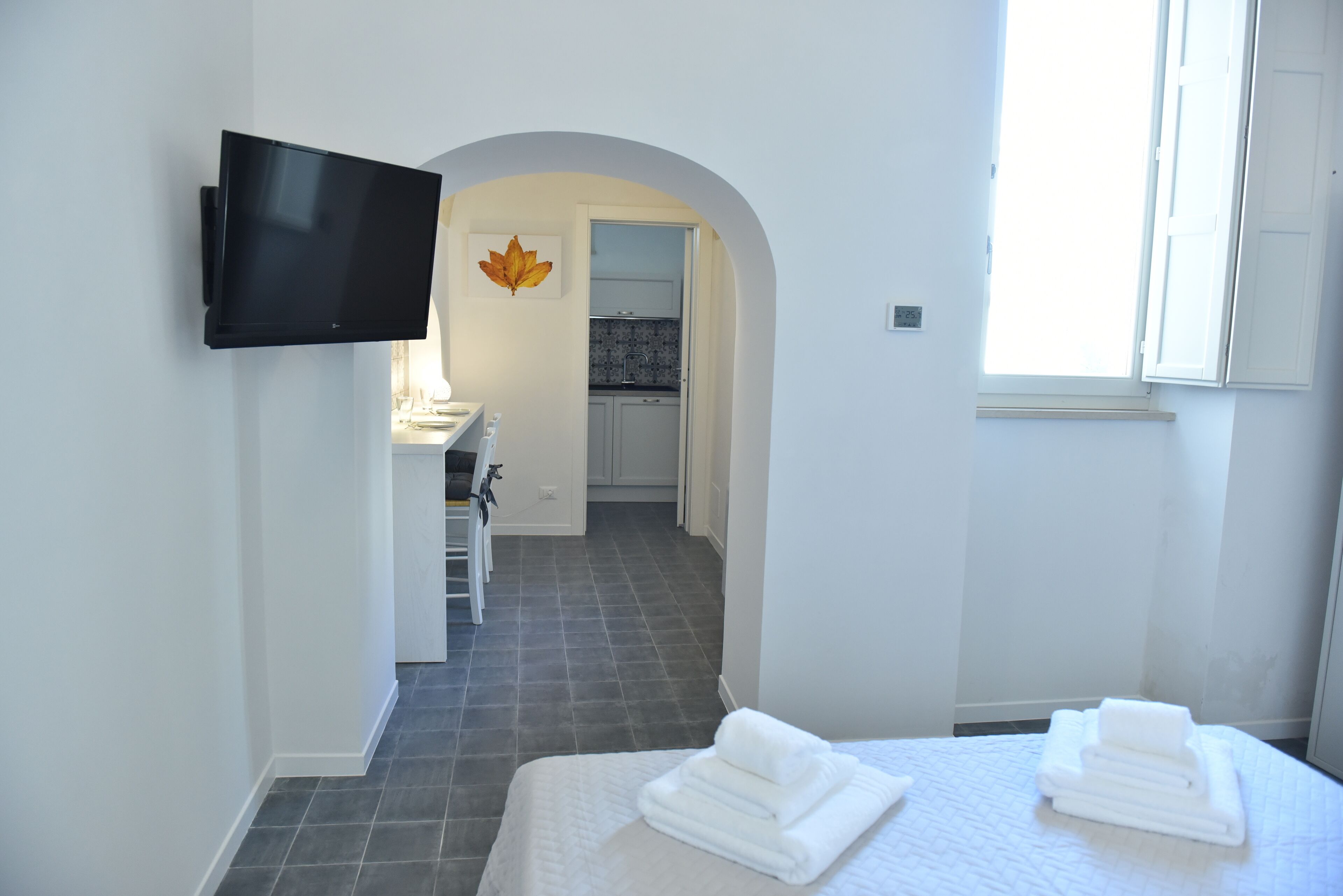 Une chambre d’hôtel avec un lit, un téléviseur fixé au mur, une salle de bain avec un lavabo et une vue sur la cuisine à travers une arche.