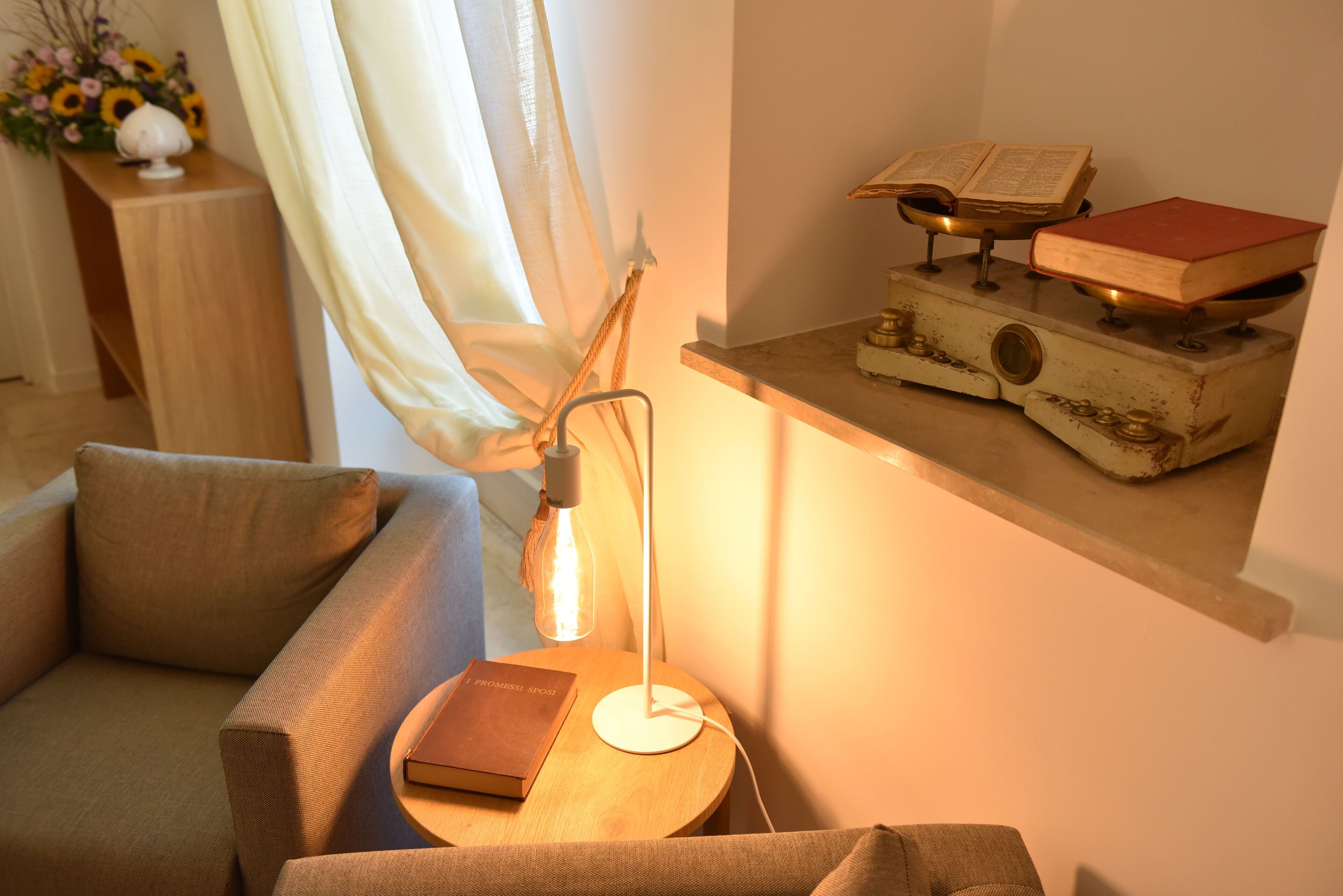 Uma sala de estar acolhedora com um sofá, uma mesinha de apoio com um livro, um abajur e uma estante com livros e um objeto decorativo.