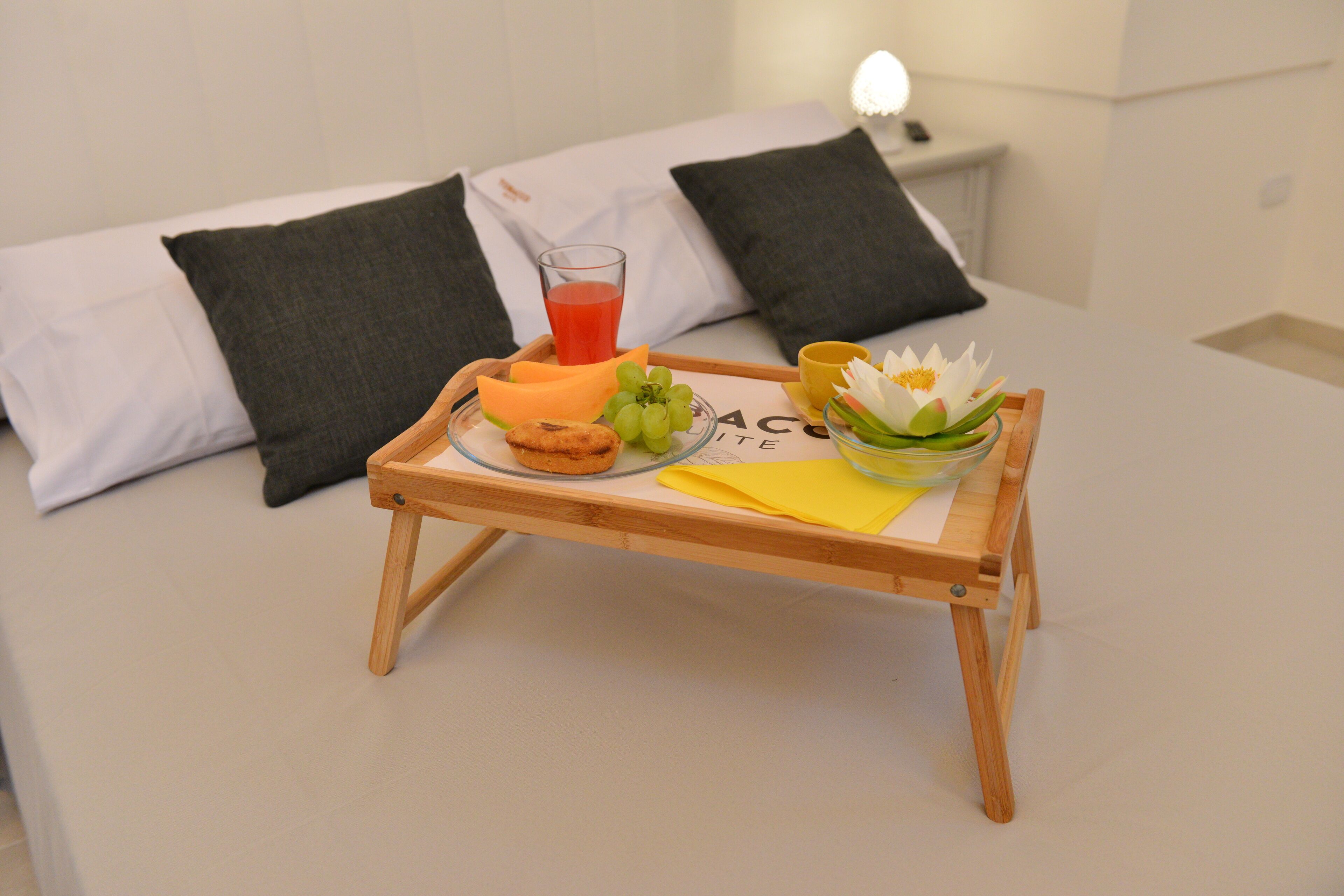 Una cama bien tendida con almohadas blancas y oscuras, una mesita de noche con una lámpara y una bandeja con alimentos para el desayuno, incluyendo tostadas, fruta y una flor.