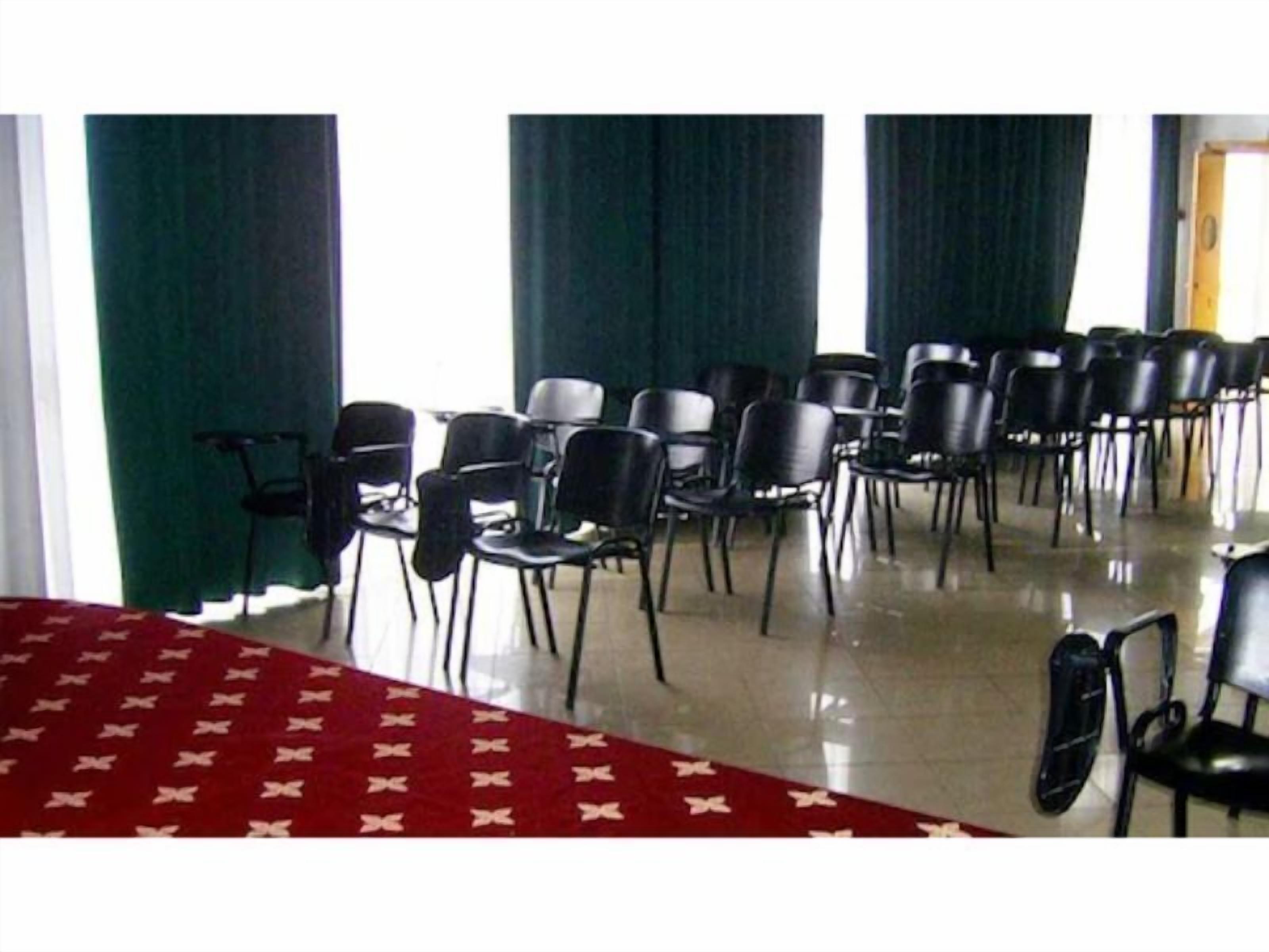 Ein Konferenzraum mit Reihen schwarzer Stühle, einem roten Teppich und grünen Vorhängen.