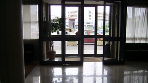 Un vestíbulo de hotel con suelo de baldosas, un mostrador de recepción y un gran ventanal con vistas a la calle y coches aparcados.