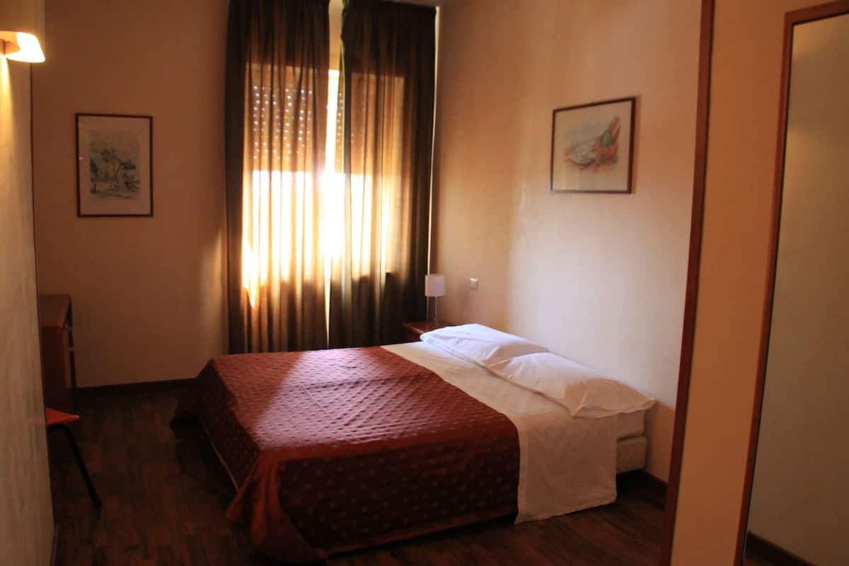 A hotel room with a bed, a chair, a desk, and a window with curtains.