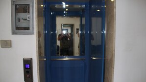 Un ascensor con pantalla digital, panel de control y espejo que refleja a una persona tomando una fotografía.