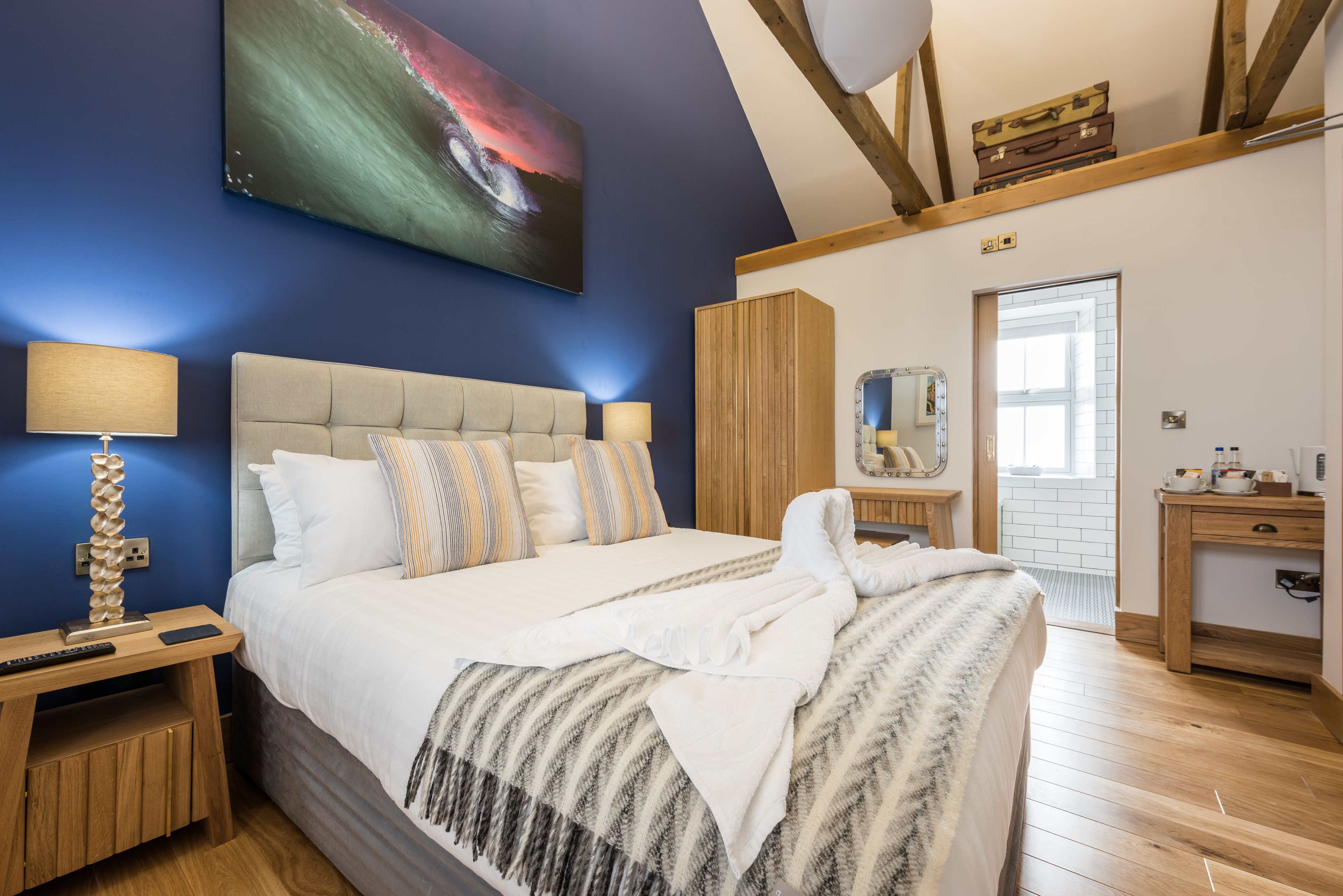 Ein modernes Schlafzimmer mit einem großen Bett, einem Nachttisch aus Holz mit einer Lampe, einer Holzkommode und einem Gemälde einer Welle an der Wand.