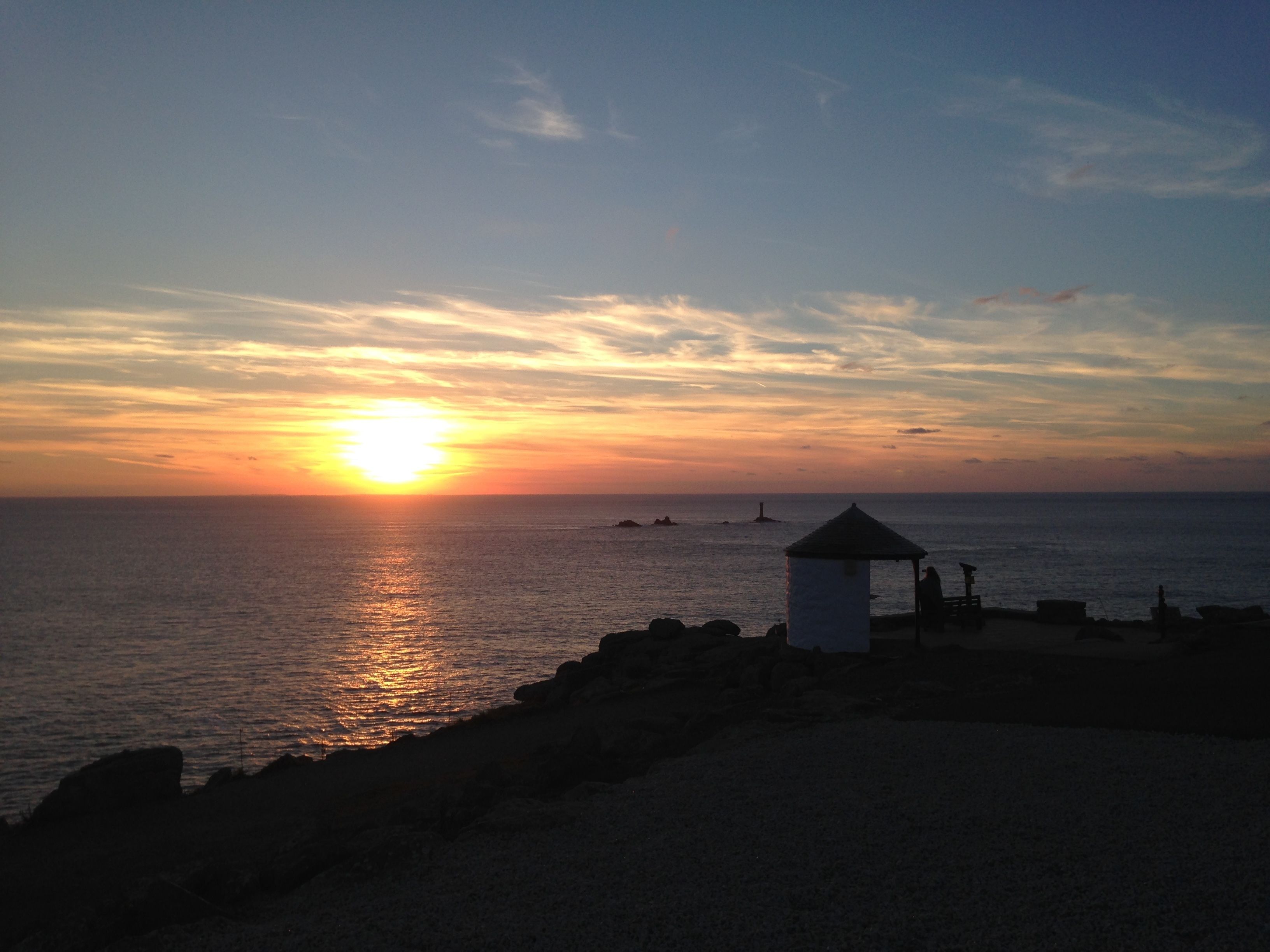 Sonnenuntergang an der Küste mit Leuchtturm und einem kleinen weißen Gebäude am Ufer.