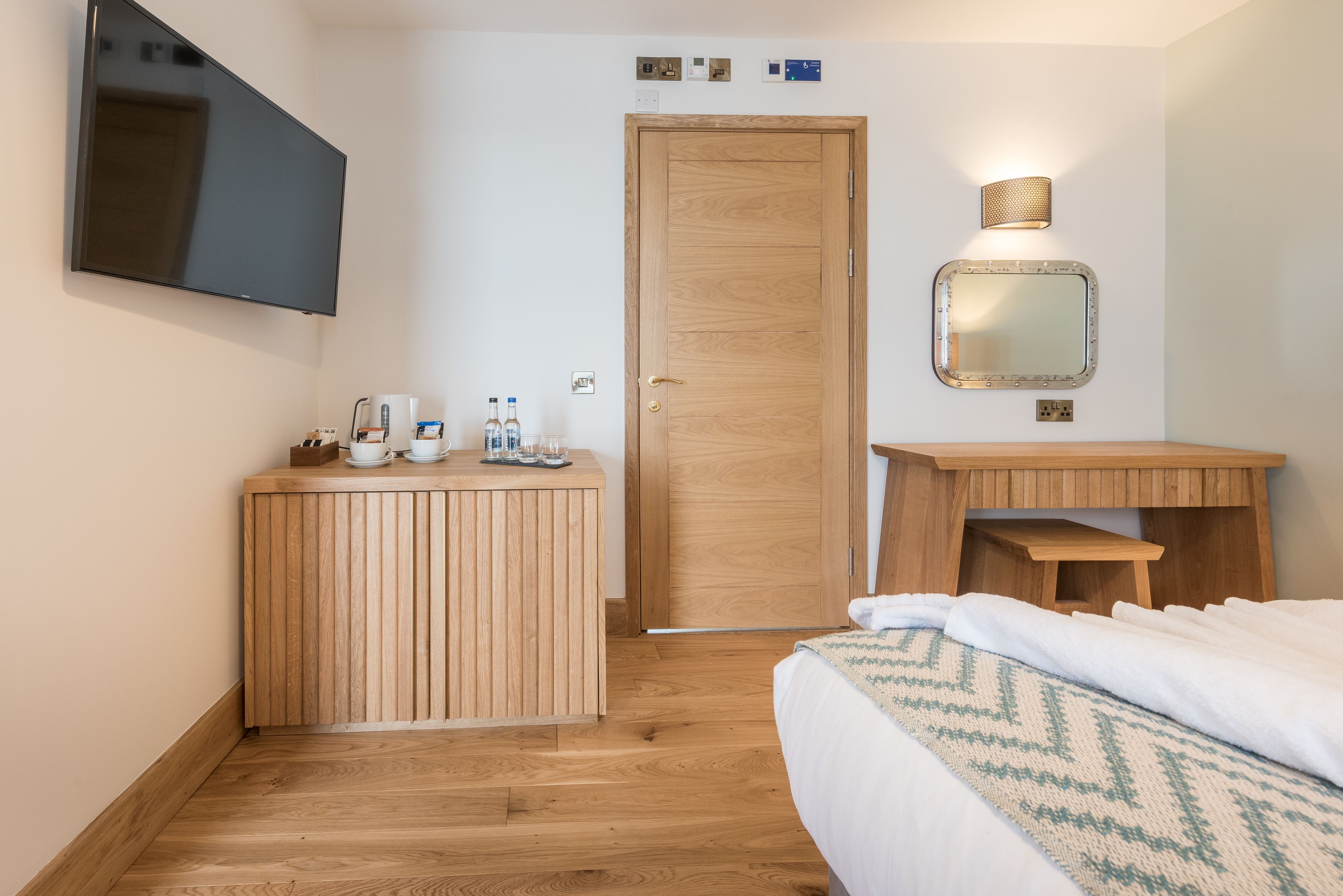 A hotel room with a wooden bed, a bedside table, a TV mounted on the wall, a wooden door, and a small wooden cabinet.