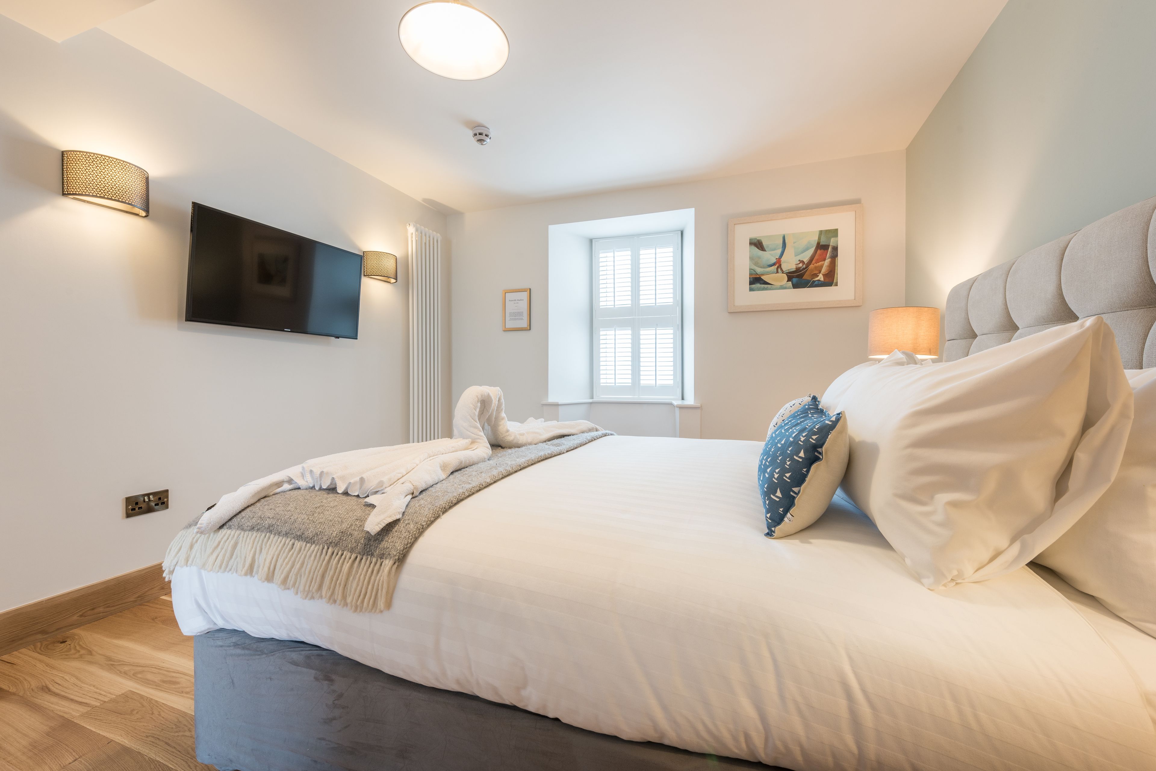 A modern bedroom with a large bed, a flat-screen TV mounted on the wall, a window with blinds, and a painting on the wall.