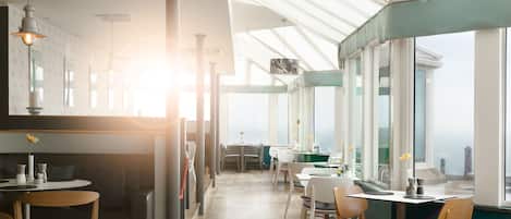 A modern restaurant with a high ceiling, wooden flooring, and large windows allowing natural light.