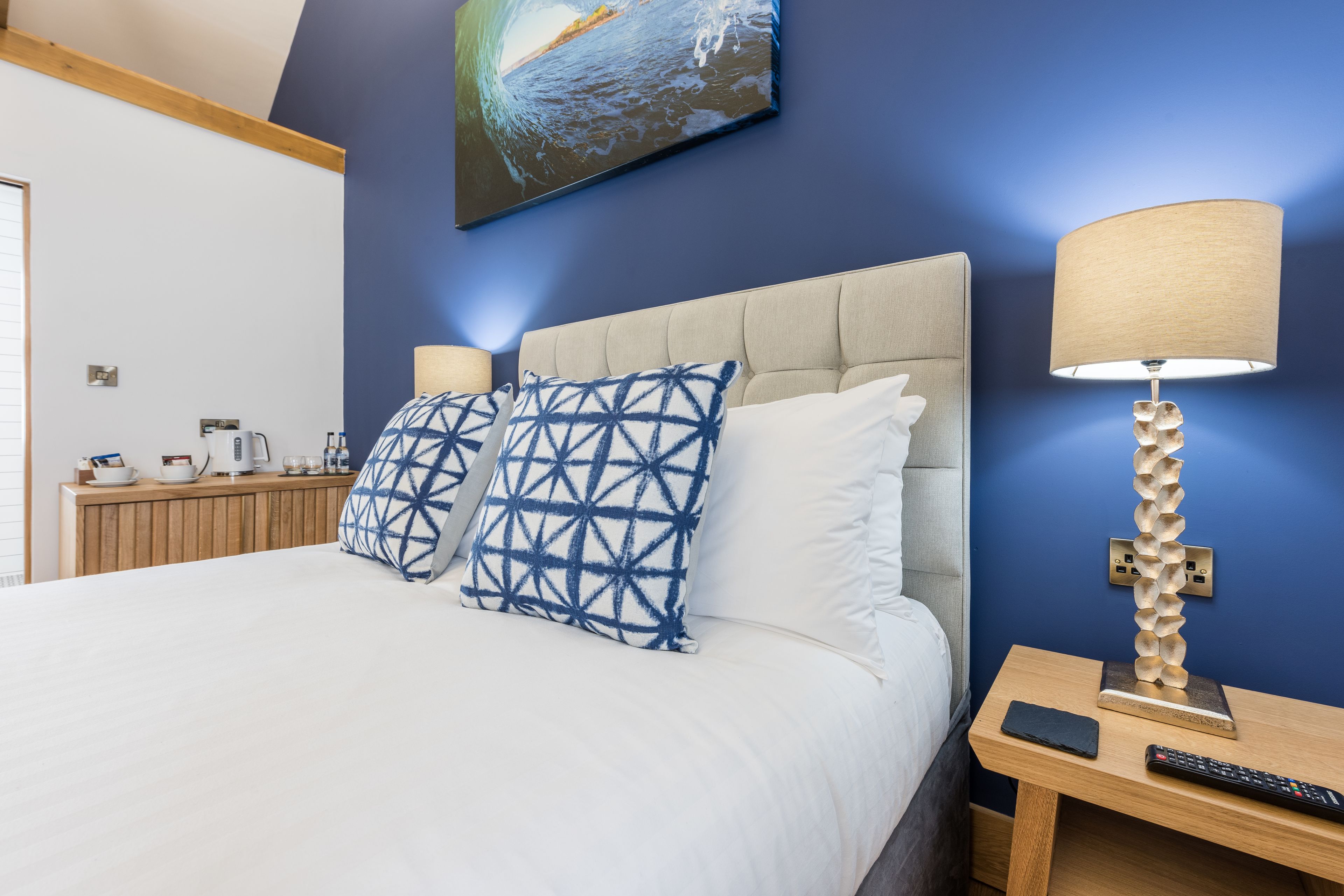 A neatly made bed with two patterned pillows, a bedside lamp, and a remote control on a wooden nightstand.