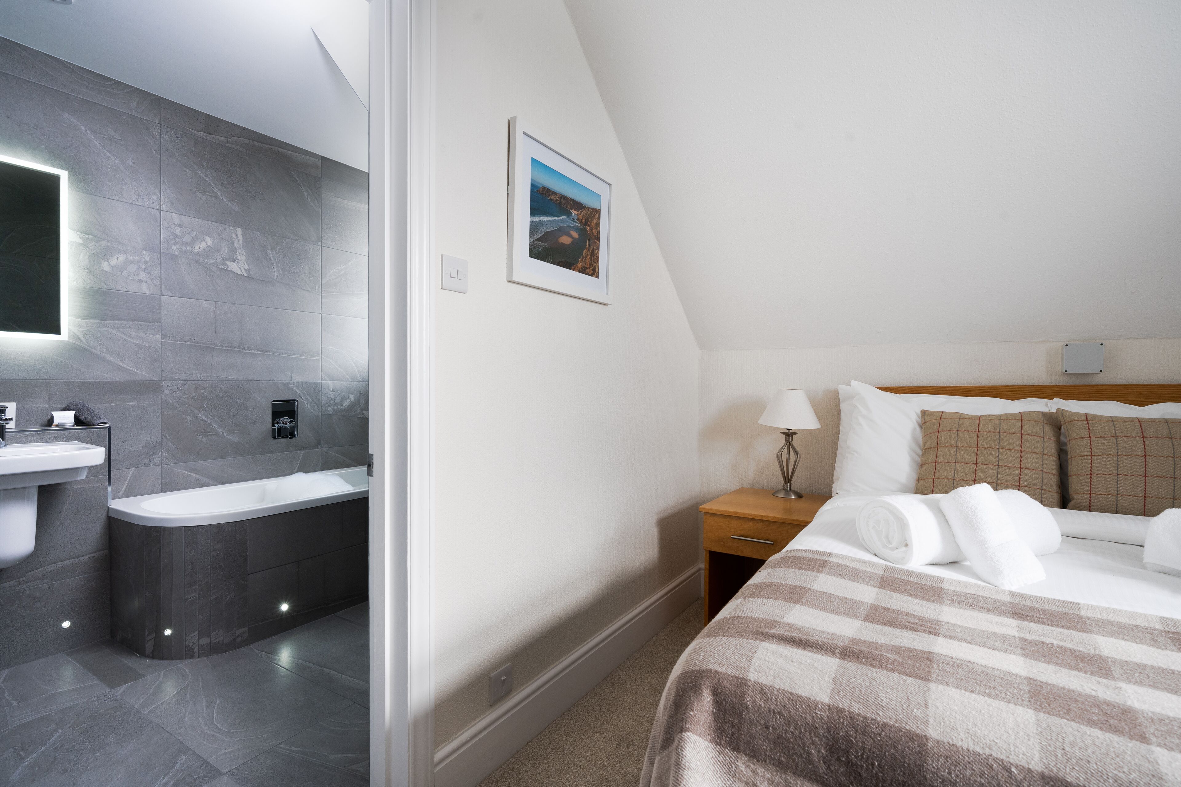 A modern hotel room with a bathroom featuring a bathtub, a sink, and a mirror. There is a bed with a checkered blanket and pillows, a bedside table with a lamp, and a framed picture on the wall.