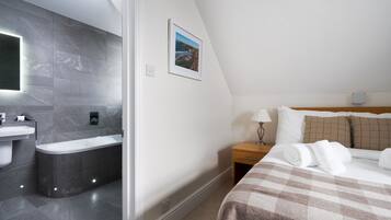A modern hotel room with a bathroom featuring a bathtub, a sink, and a mirror. There is a bed with a checkered blanket and pillows, a bedside table with a lamp, and a framed picture on the wall.