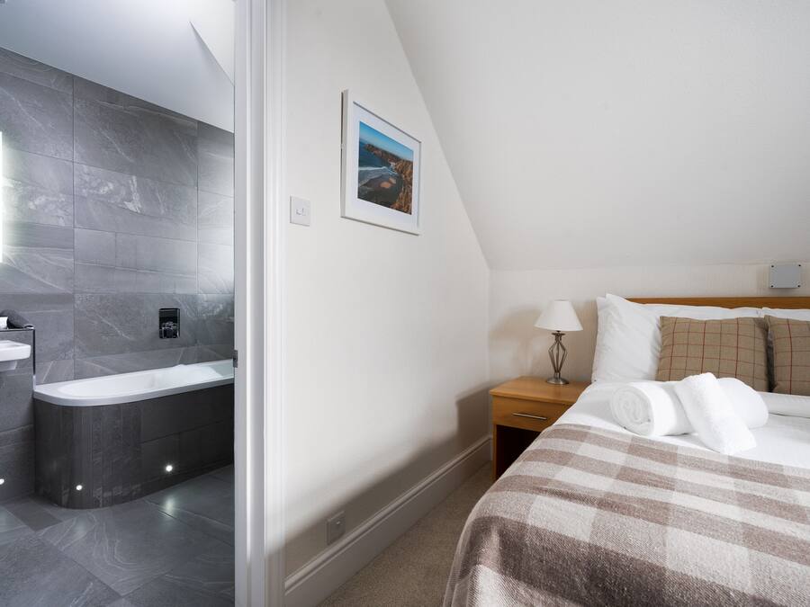 A modern hotel room with a bathroom featuring a bathtub, a sink, and a mirror. There is a bed with a checkered blanket and pillows, a bedside table with a lamp, and a framed picture on the wall.