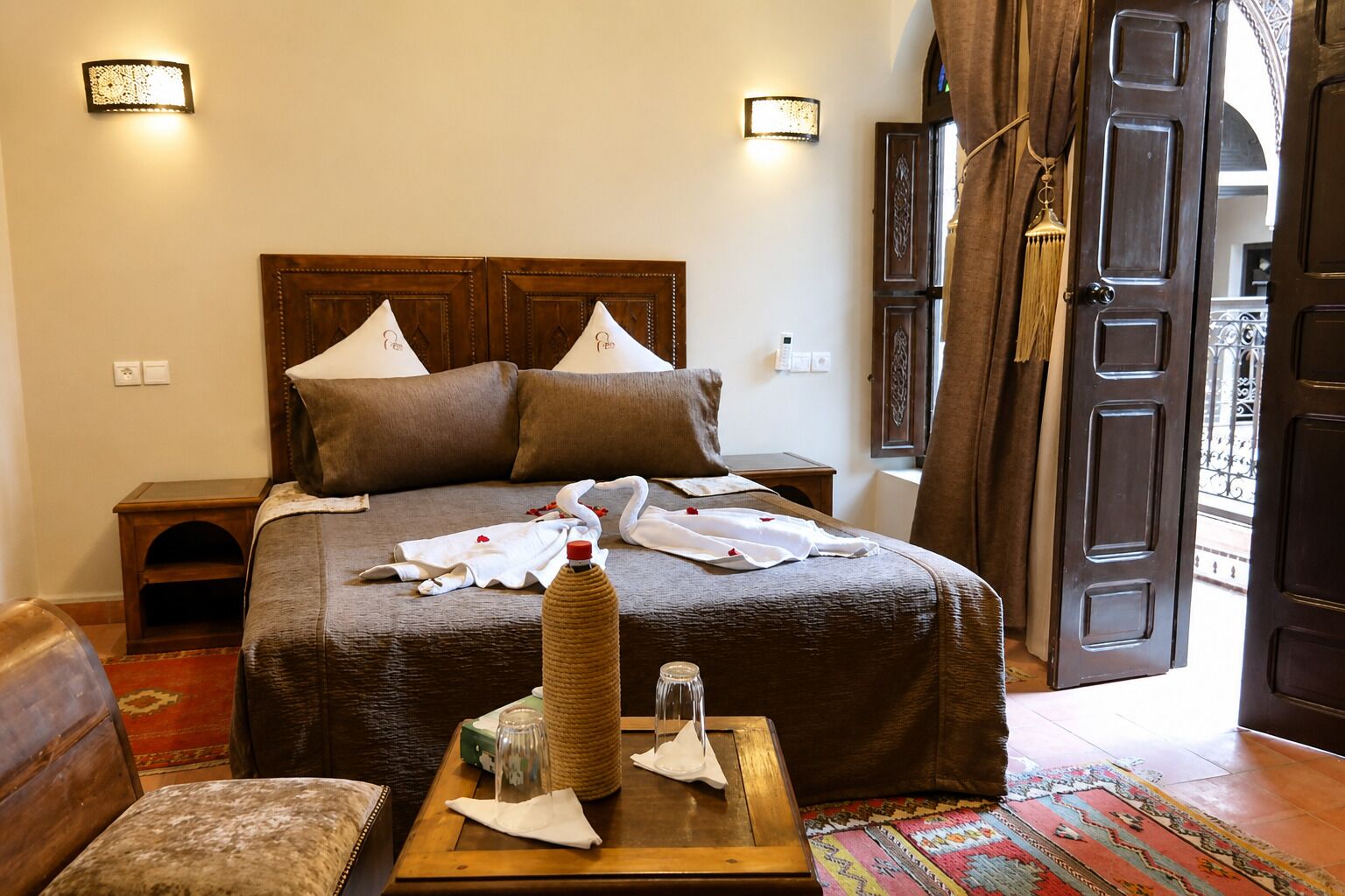 A neatly made bed with pillows, a bedside table with a bottle and glass, a chair, and a colorful rug.