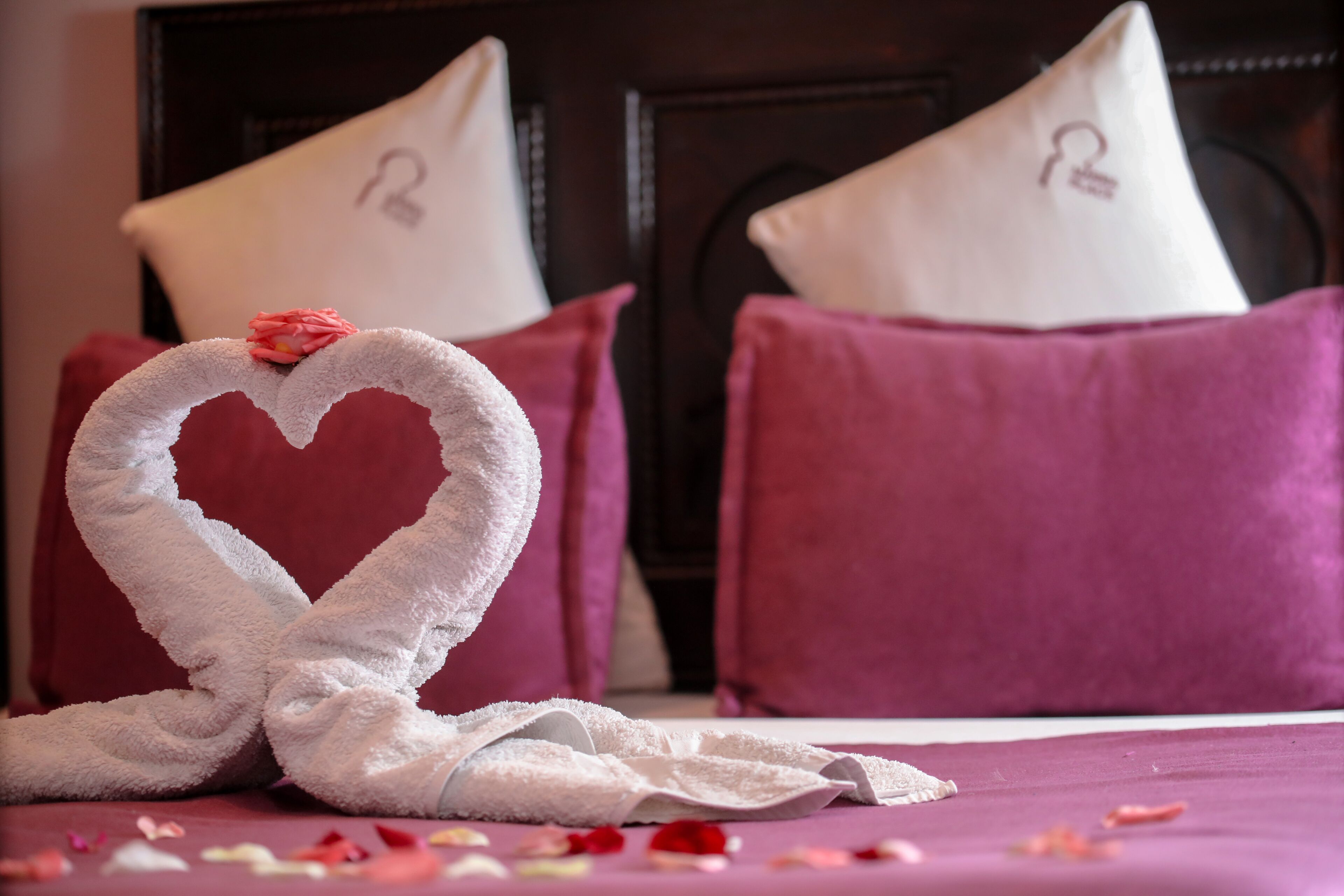A bed with a heart-shaped towel and rose petals, surrounded by purple pillows and a dark headboard.