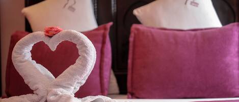 A bed with a heart-shaped towel and rose petals, surrounded by purple pillows and a dark headboard.