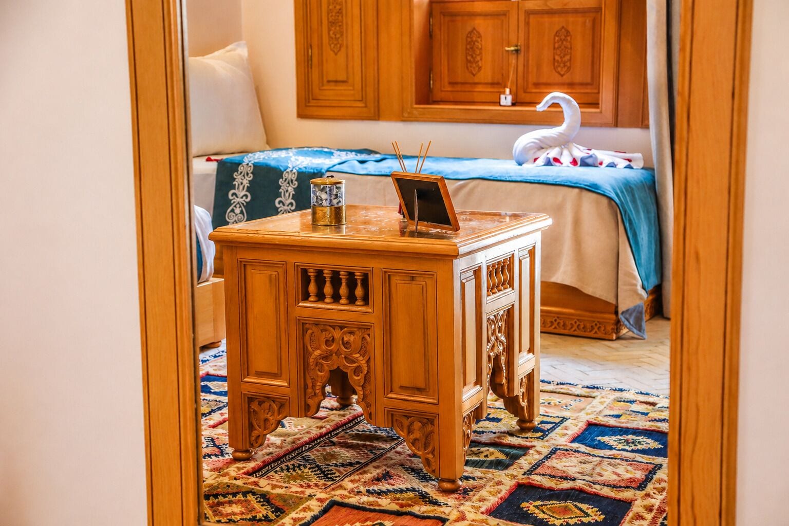 A room with a bed, a wooden side table with a laptop, a decorative swan, and a patterned carpet.