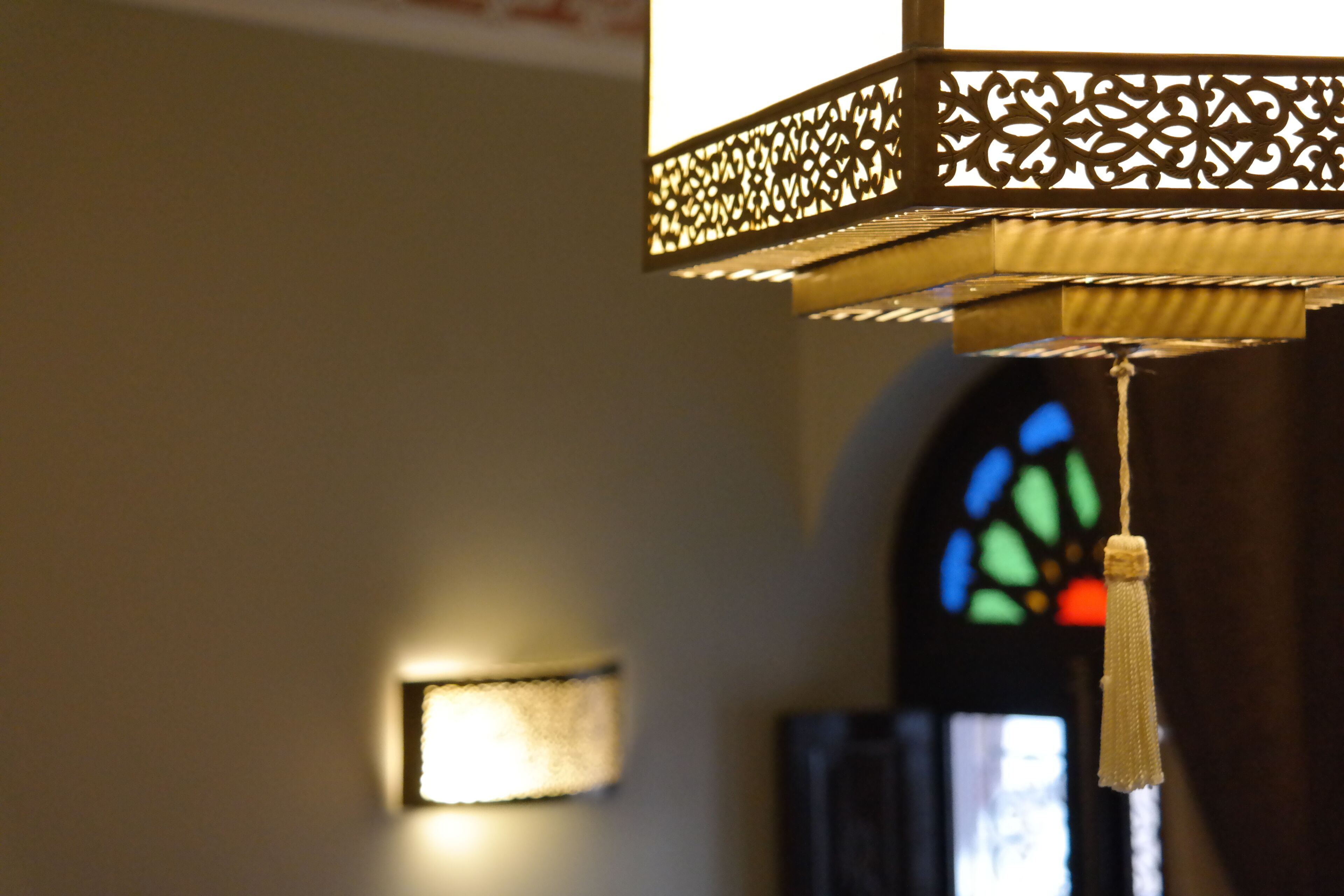 Un luminaire suspendu orné d’un motif complexe en treillis, un applique murale et un vitrail en arrière-plan.