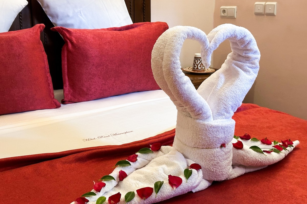 A hotel room with a bed covered in a red blanket, pillows, and a towel arranged in a heart shape with rose petals.
