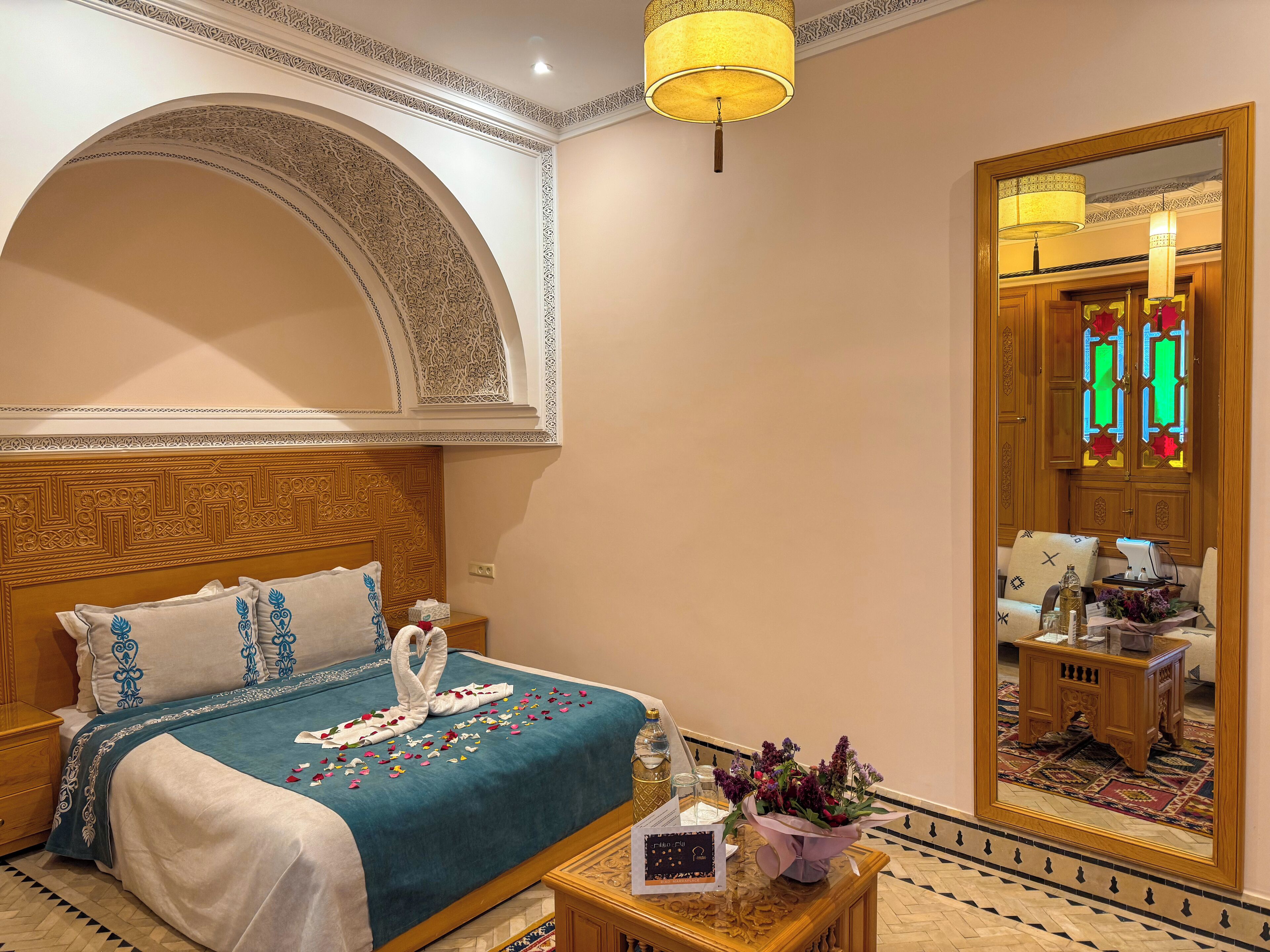 A hotel room with a large bed, a wooden headboard, a small table with a vase of flowers, and a mirror reflecting a door with stained glass.